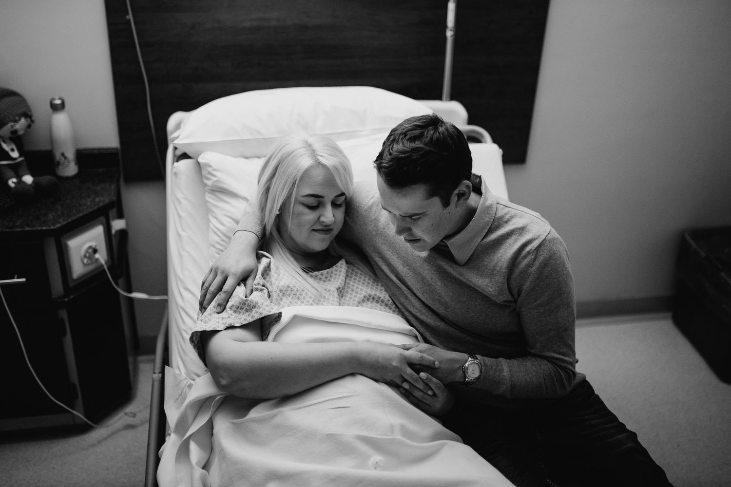 Black and white photo of new parents in hospital room, mother in bed holding newborn while father leans in closely.
