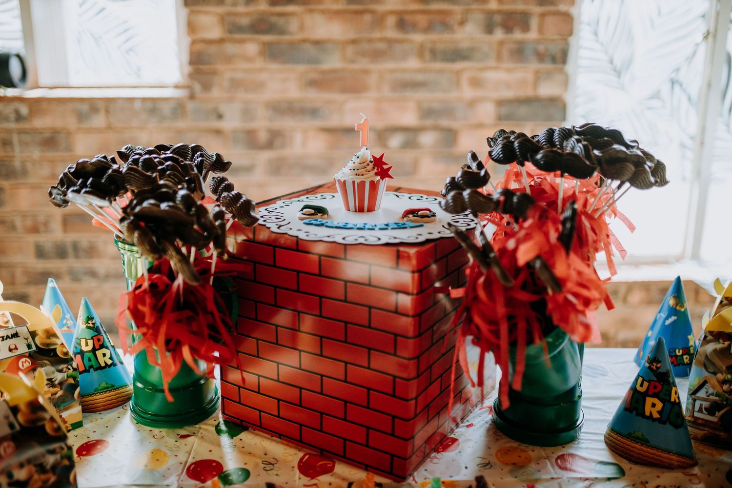 Pink and turquoise baby shower decorations displayed on a rustic brick wall with string lights and balloons.
