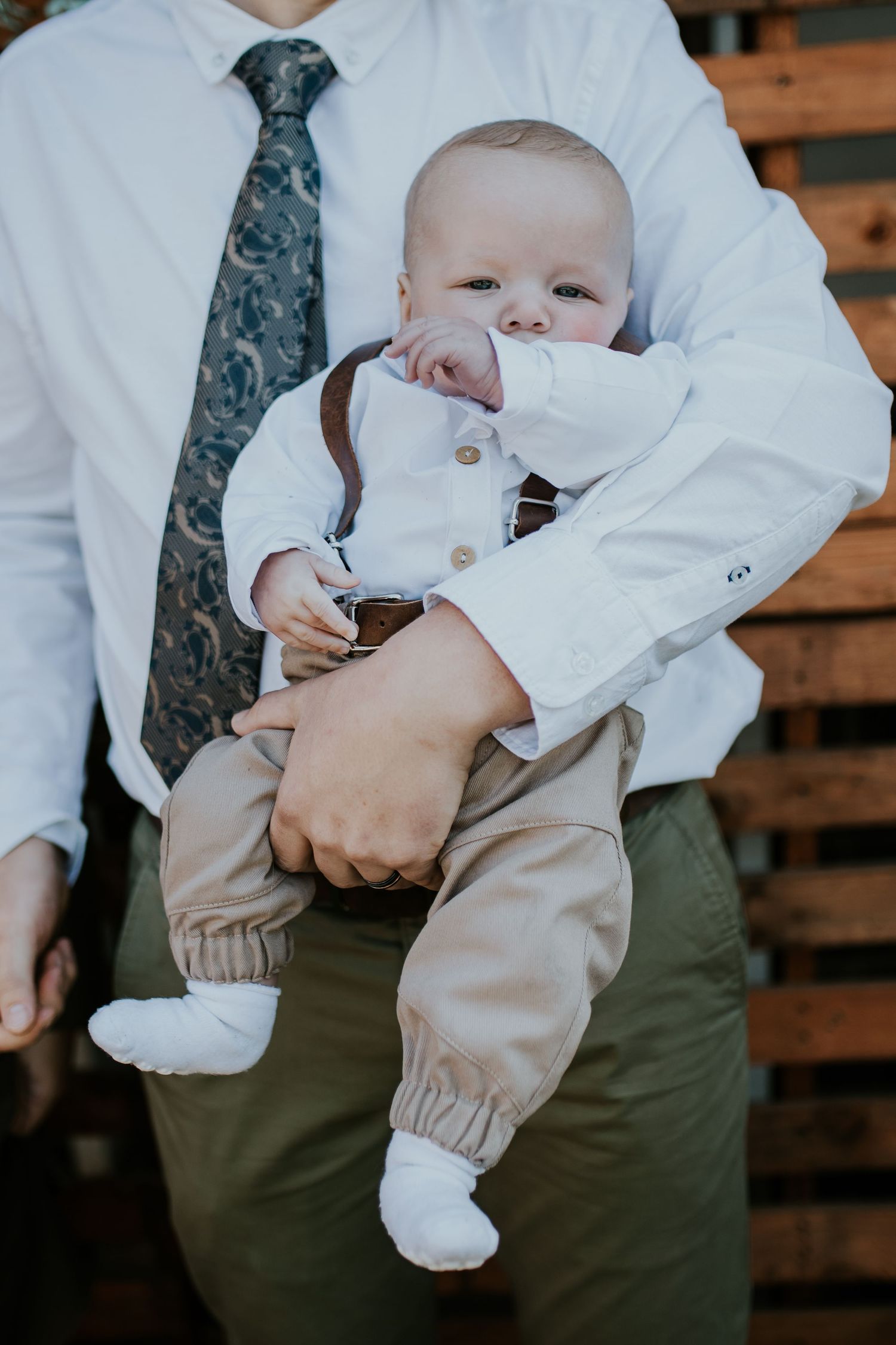 Baby wearing white button-down shirt and beige pants is held against someone in formal attire with tie and dress shirt.