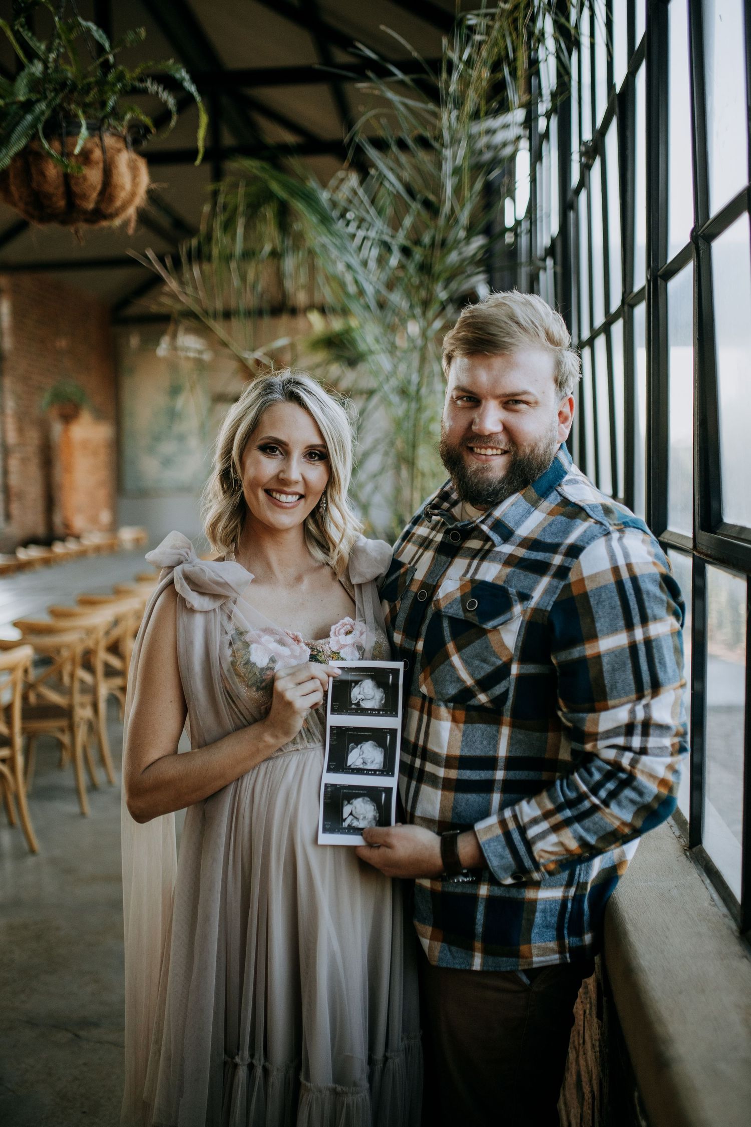 A couple stands together holding up an ultrasound photo in an indoor space with natural light.