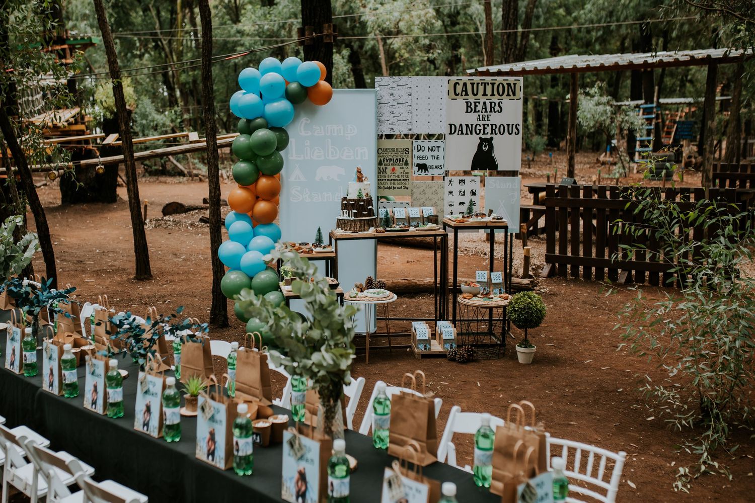 Outdoor wedding ceremony setting with blue and white balloons decorating a forest venue featuring wooden benches and signage.