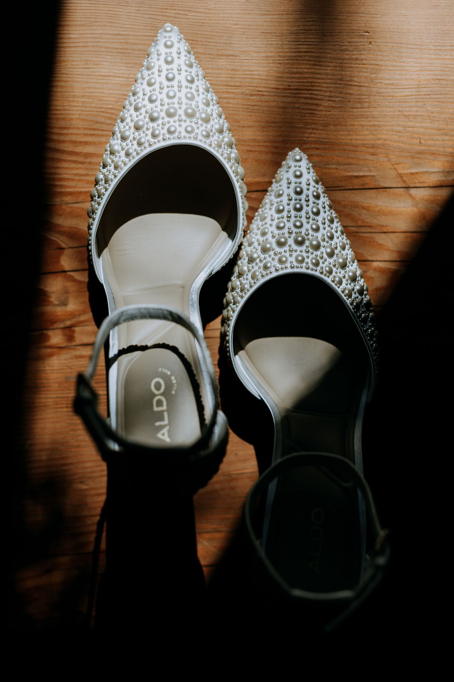 Elegant bridal shoes with crystal embellished pointed toes and ankle straps cast dramatic shadows on wooden floor.