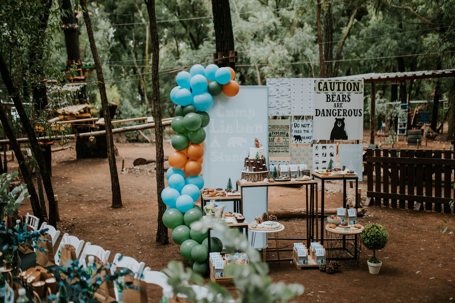 Outdoor rustic wedding venue decorated with teal and green balloon arches and wooden signs in a forest setting.