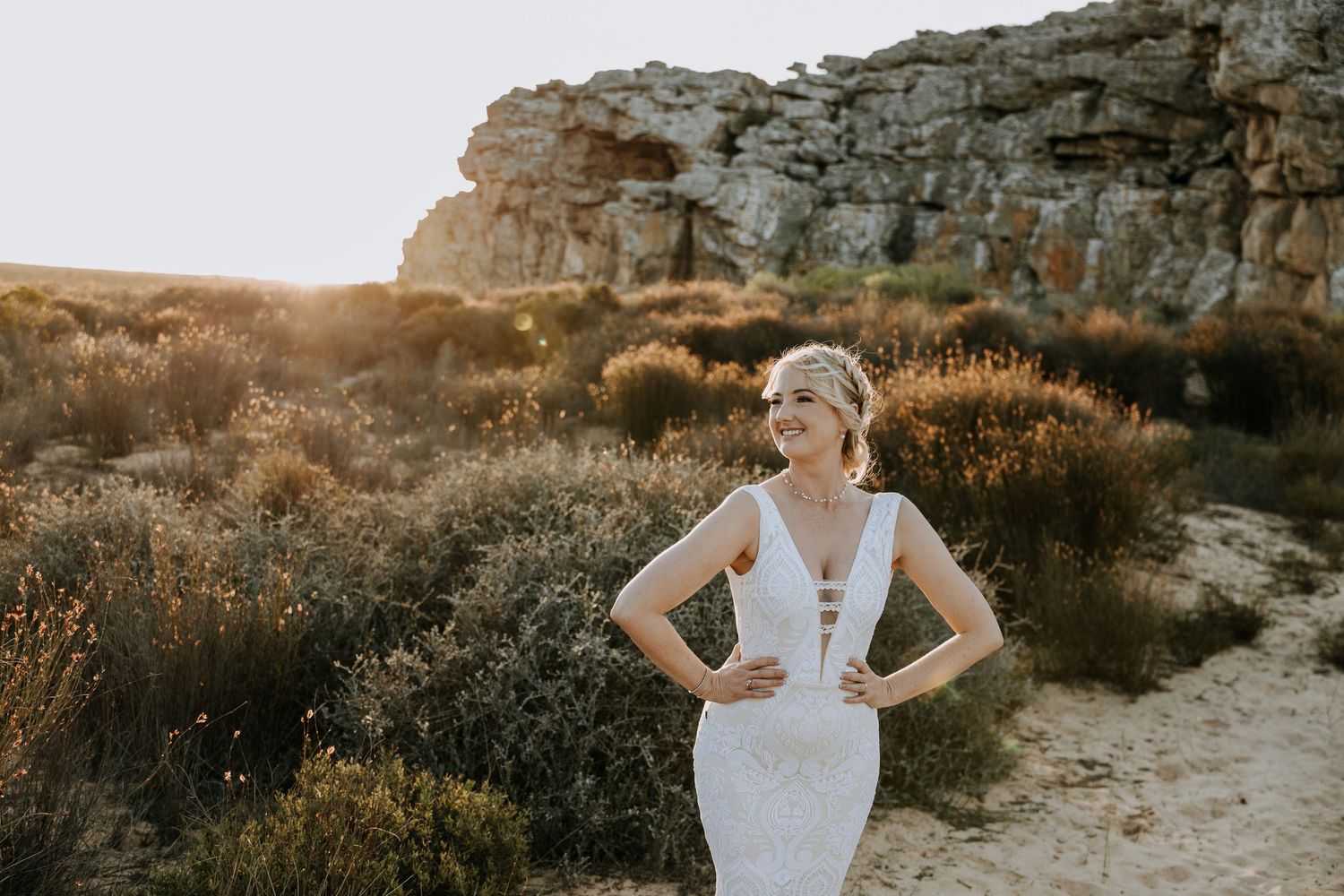 Bride in elegant lace white wedding dress poses amidst scenic rock formations at golden sunset.