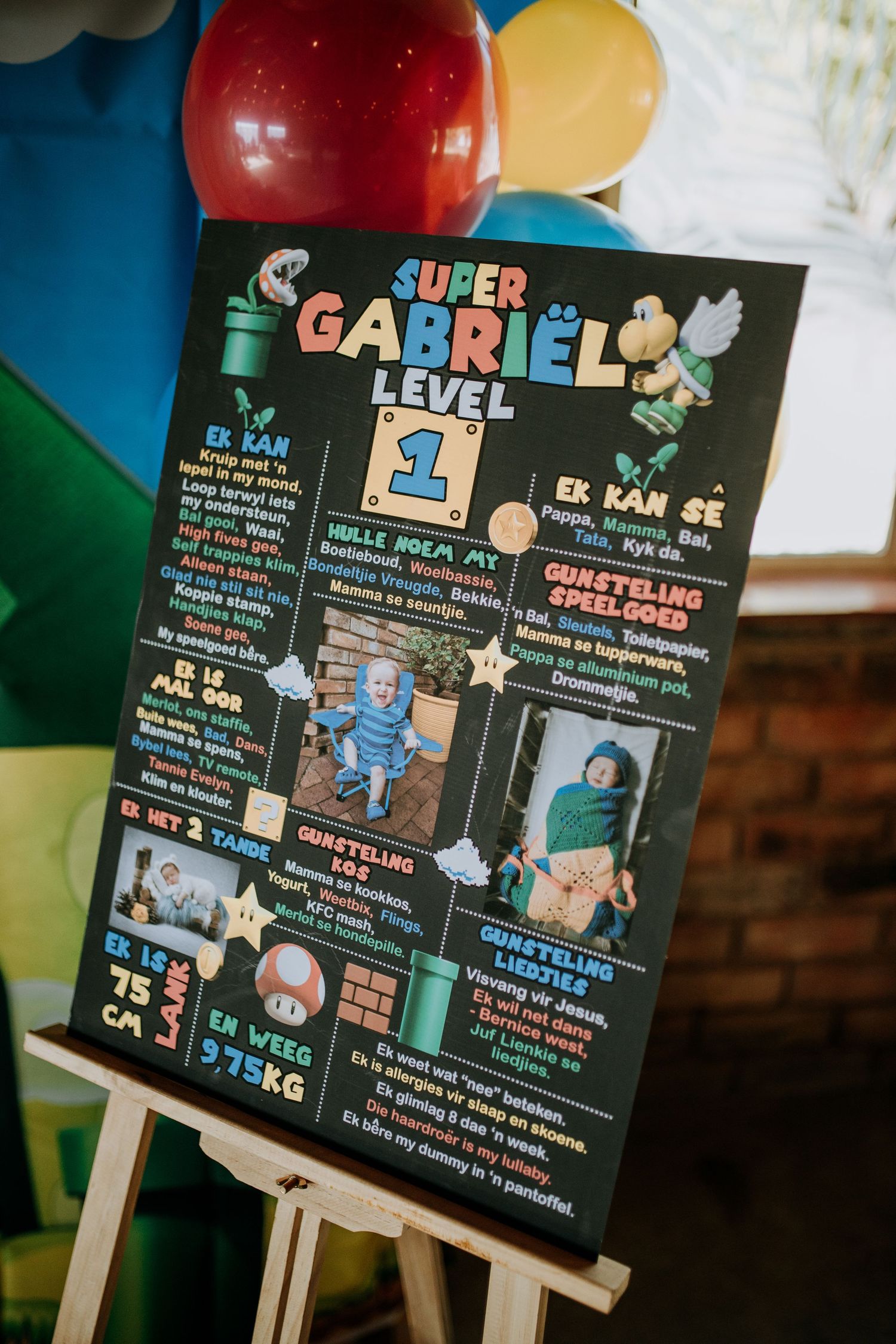 Three black chalkboard signs with Super Mario themed first birthday party milestones display on wooden easels at a celebration.