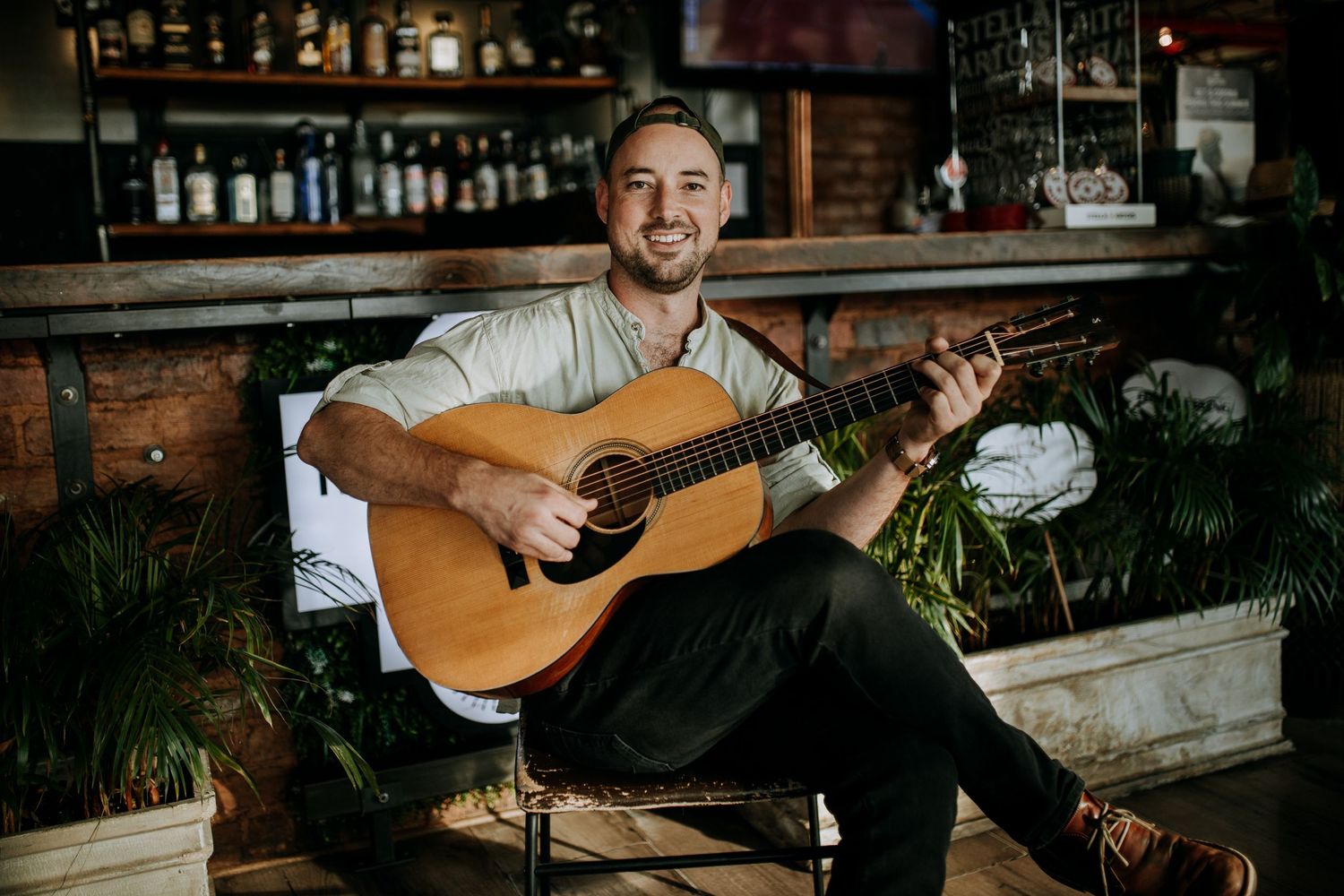 A musician performs on acoustic guitar against a rustic brick wall backdrop in a dimly lit venue with a bar setting.