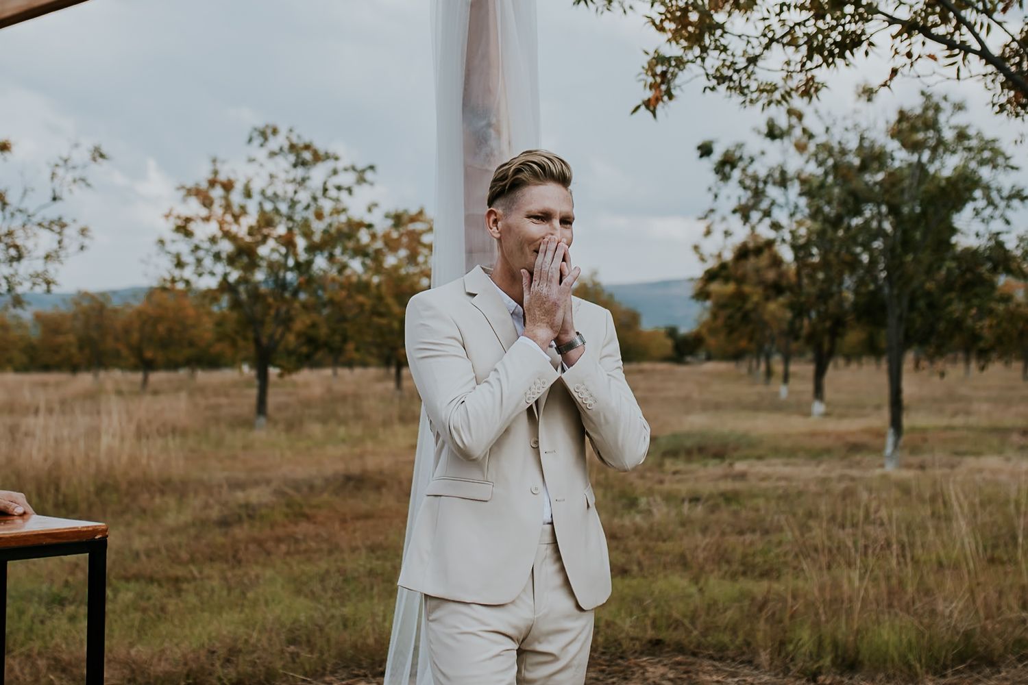 Person at a countryside wedding reacting emotionally in their cream-colored suit jacket against a rustic outdoor backdrop.