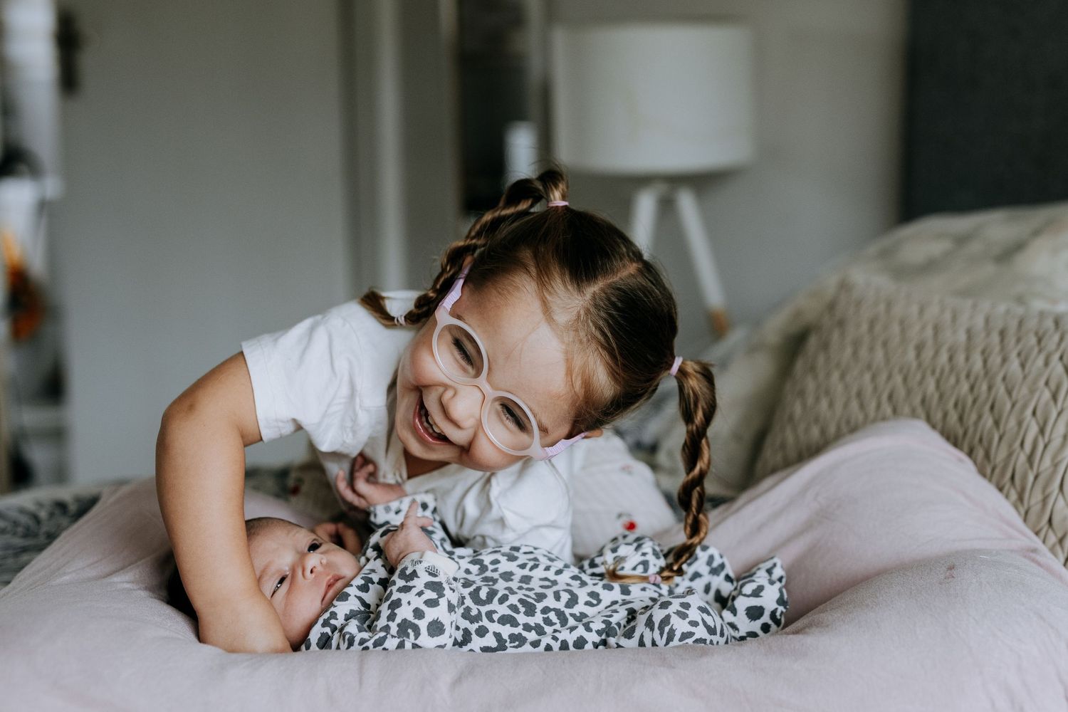 Smiling girl with braids and pink glasses leaning over a newborn baby on a bed.