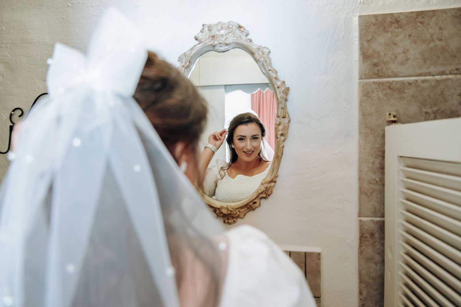 A bride in a white wedding dress and veil looks at her reflection in an ornate gold-framed mirror.