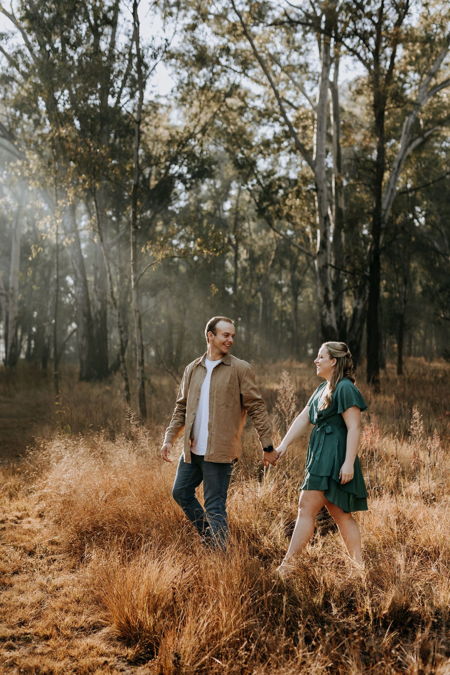 A casually dressed couple walks together through tall golden grass in a sunlit wooded area during autumn.