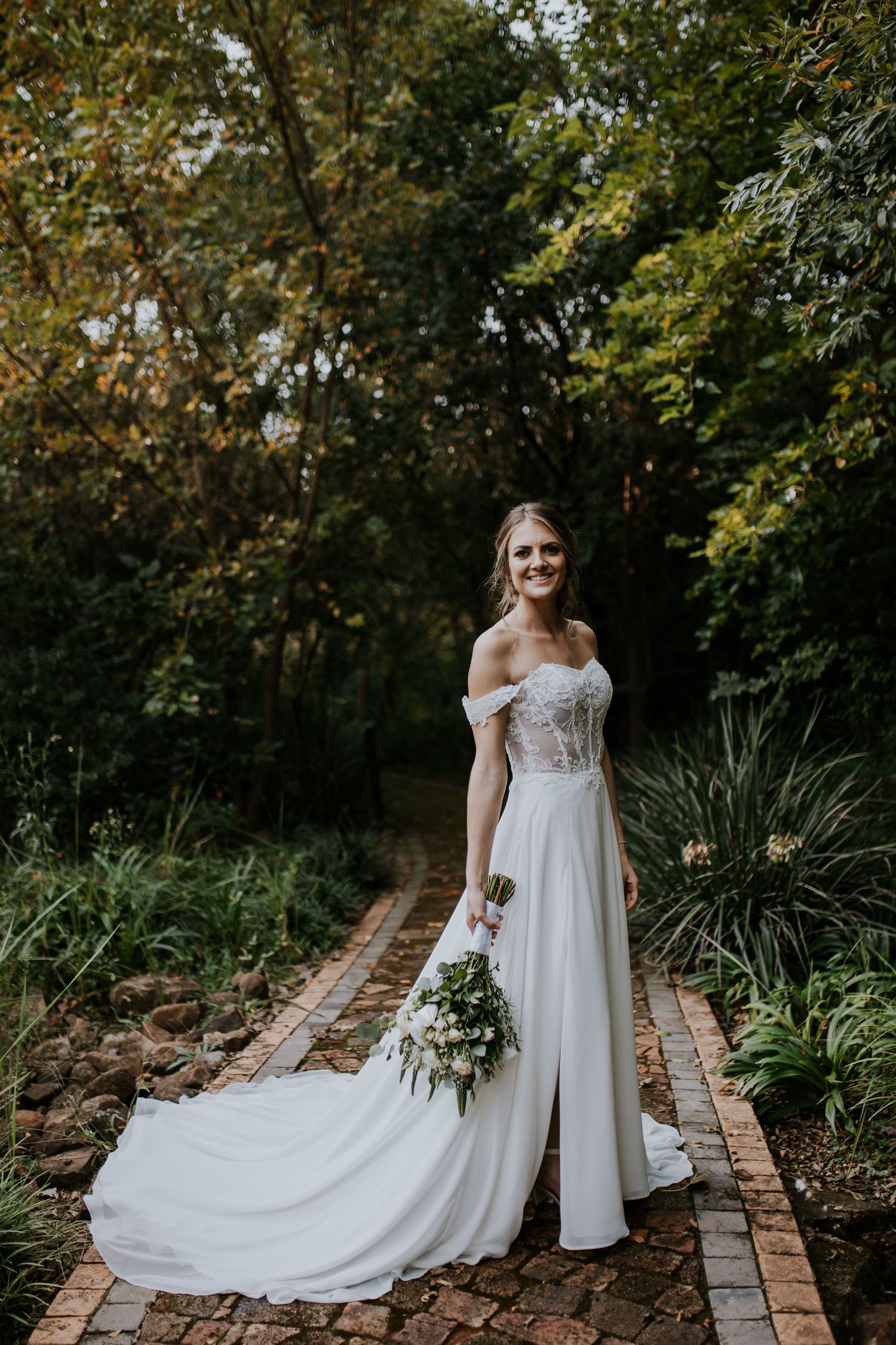 Four wedding photos showing a bride in a flowing white dress standing on a brick pathway surrounded by lush garden greenery.