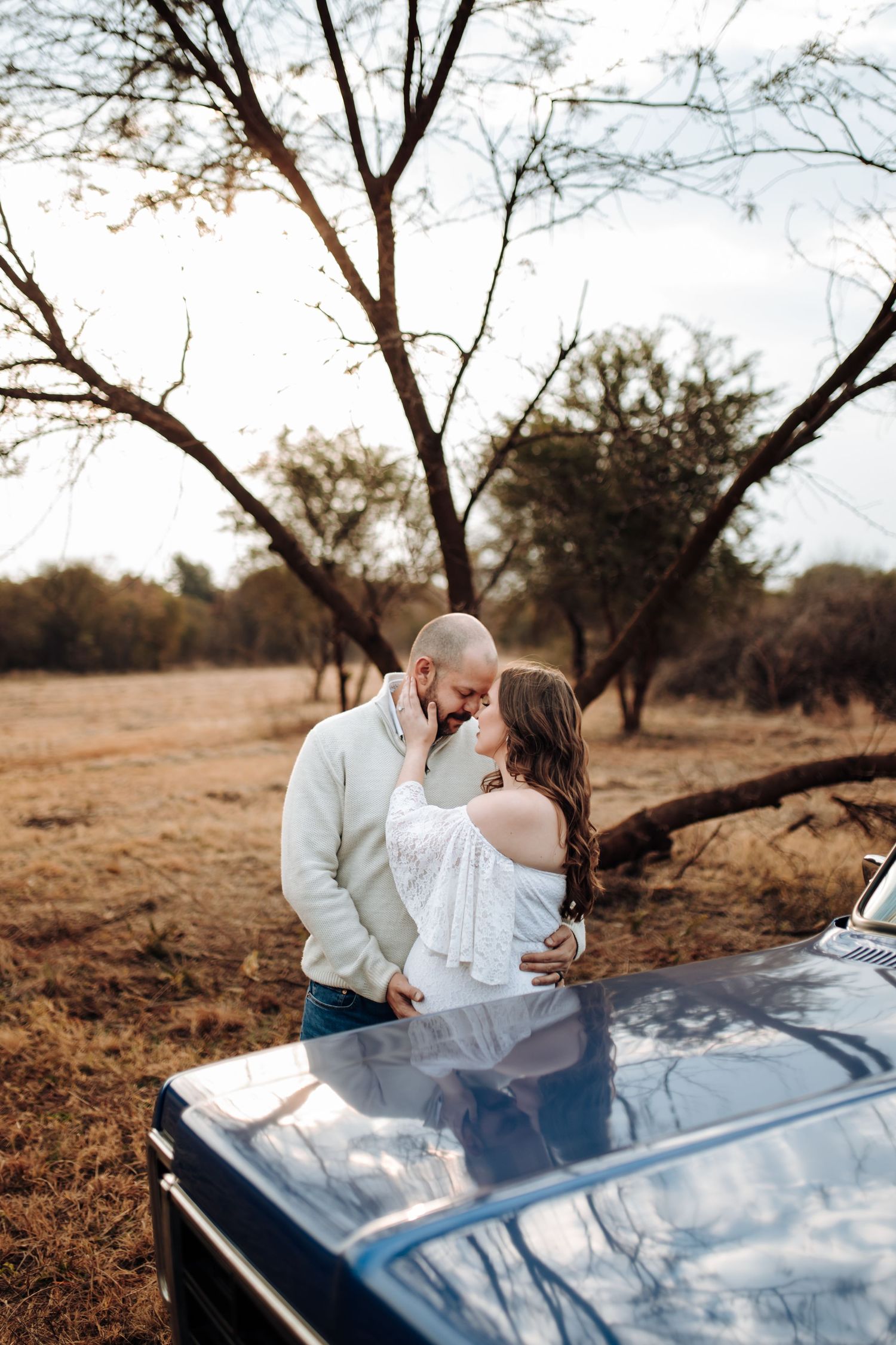 A couple shares a romantic moment leaning against a vintage car in a golden field with bare trees at sunset.