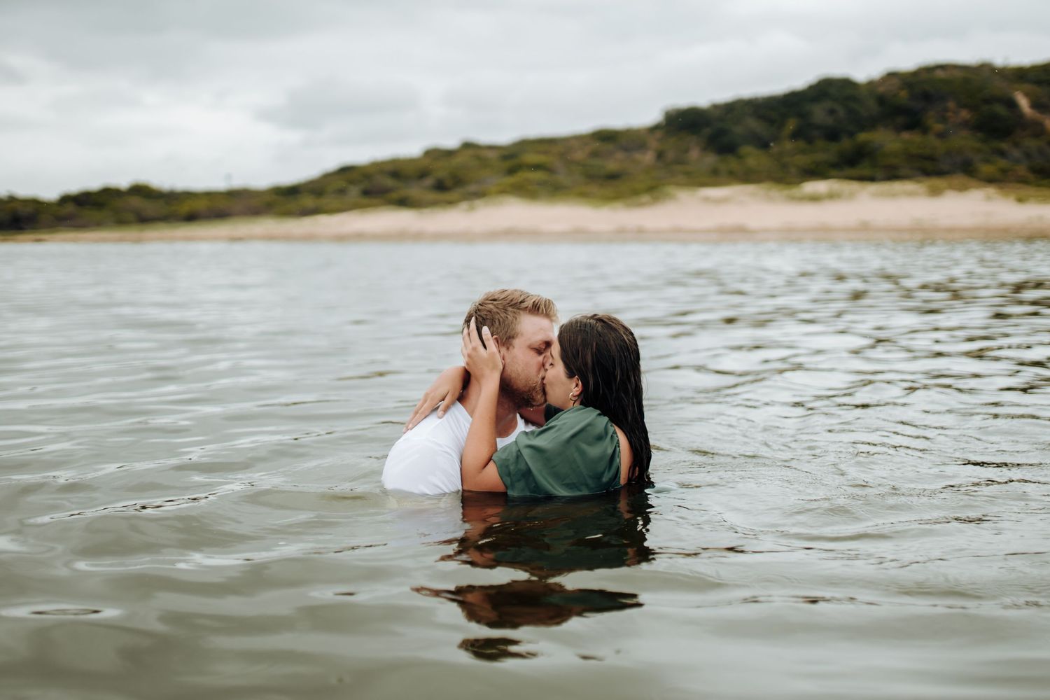 Two people share a romantic moment while wading in shallow coastal waters near a sandy beach and hillside.