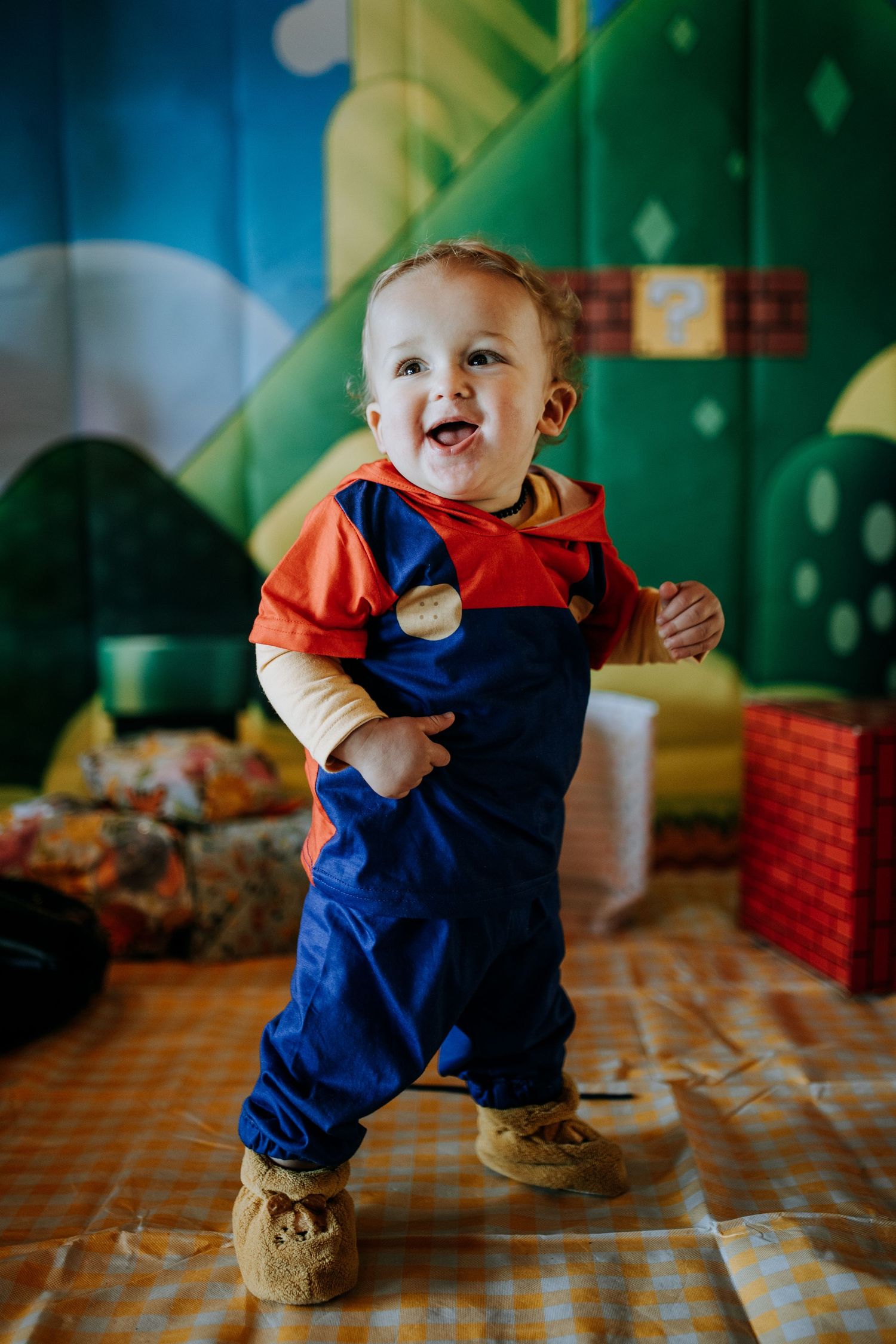 A small child wearing a Mario-inspired costume of blue overalls and red shirt laughs while standing against a colorful background.