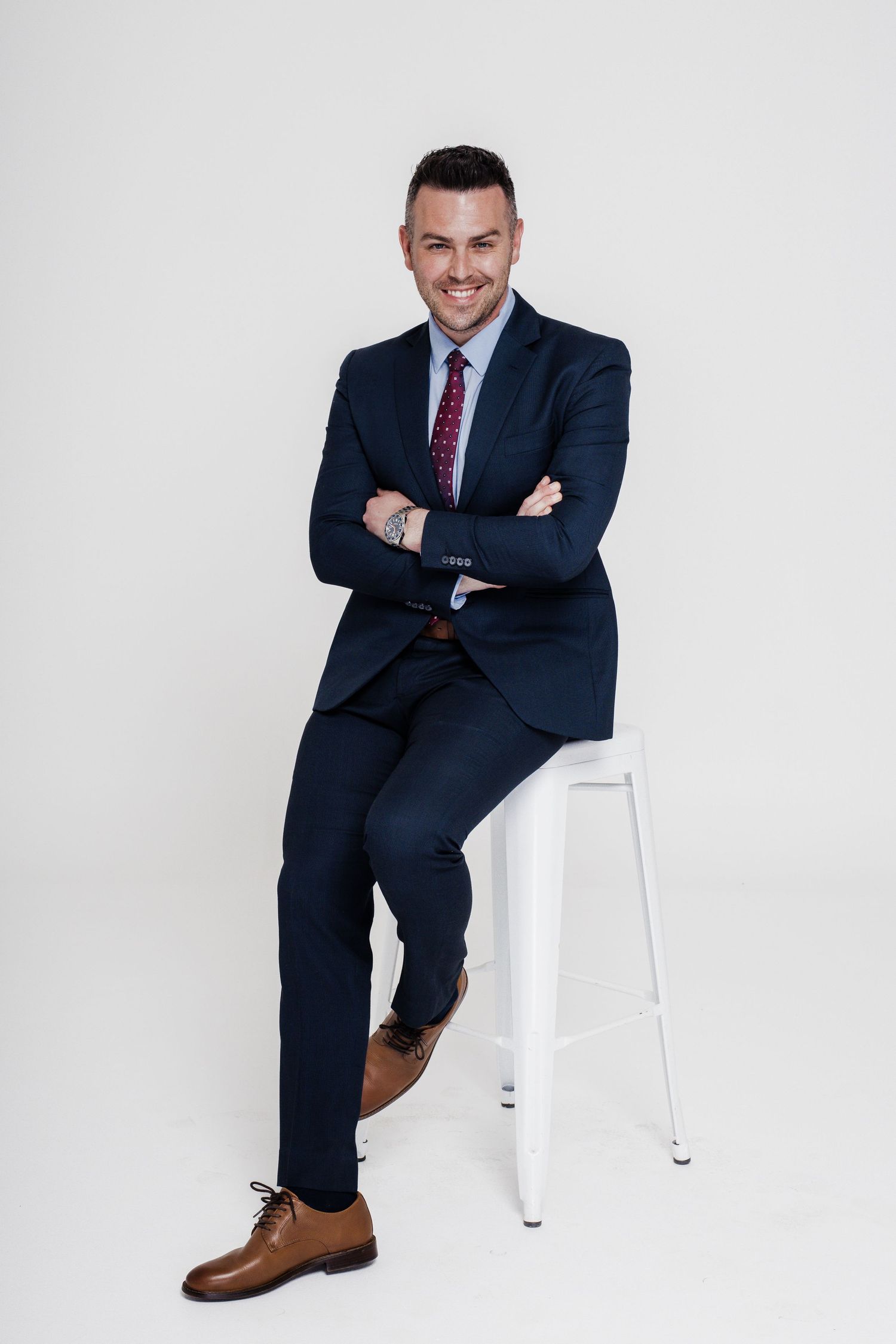 Professional in navy suit and burgundy tie sits confidently on white stool against light background.