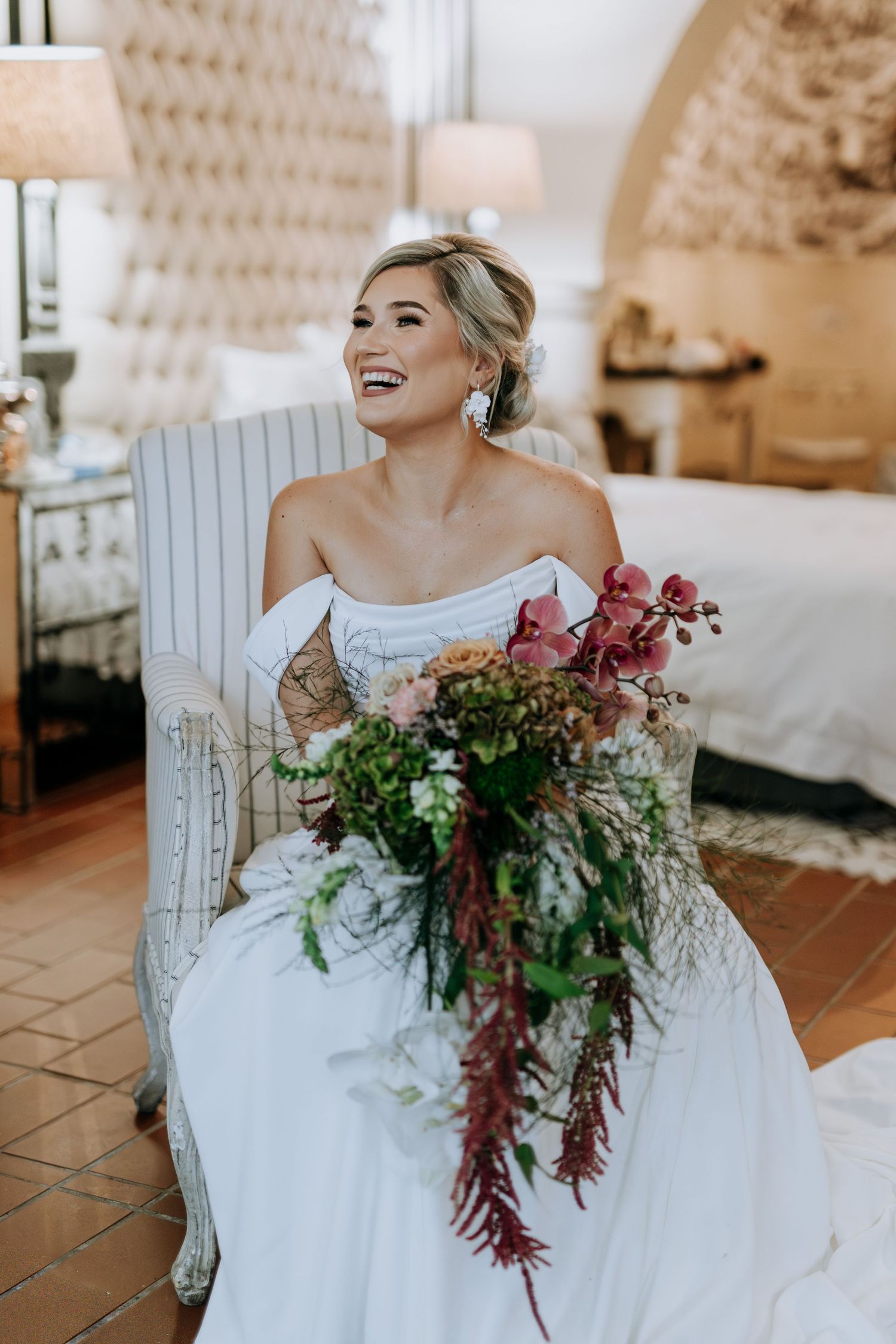 A bride in a white wedding dress sits in a vintage chair holding a cascading burgundy and green floral bouquet.