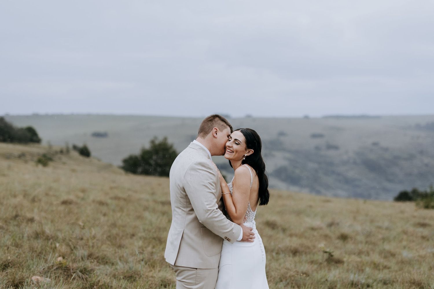 A couple shares a romantic kiss on a scenic hilltop overlooking rolling hills and mountains in the distance.