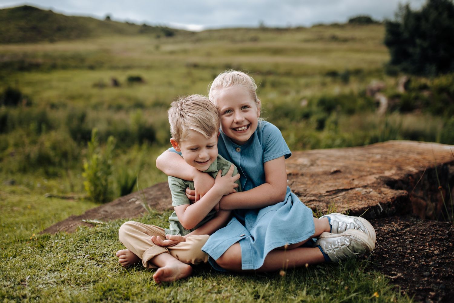Two children sit together outdoors laughing and hugging on a grassy hill with countryside landscape in the background.