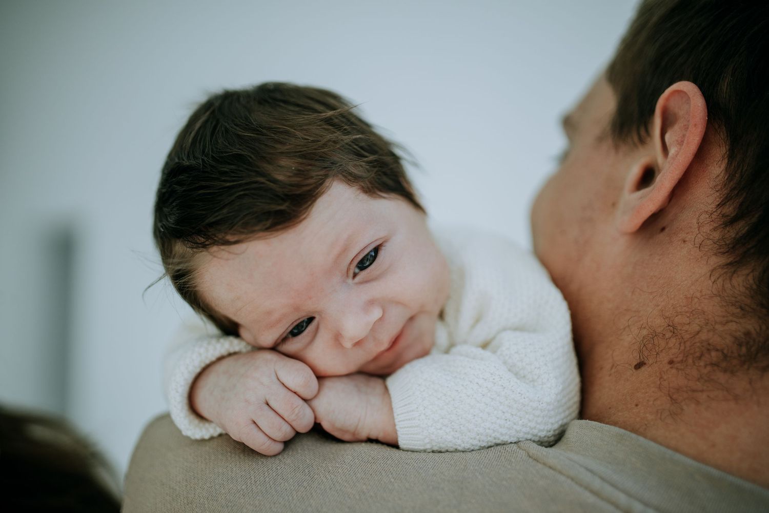 Baby in cream colored sweater rests tenderly against parent's shoulder in intimate moment.