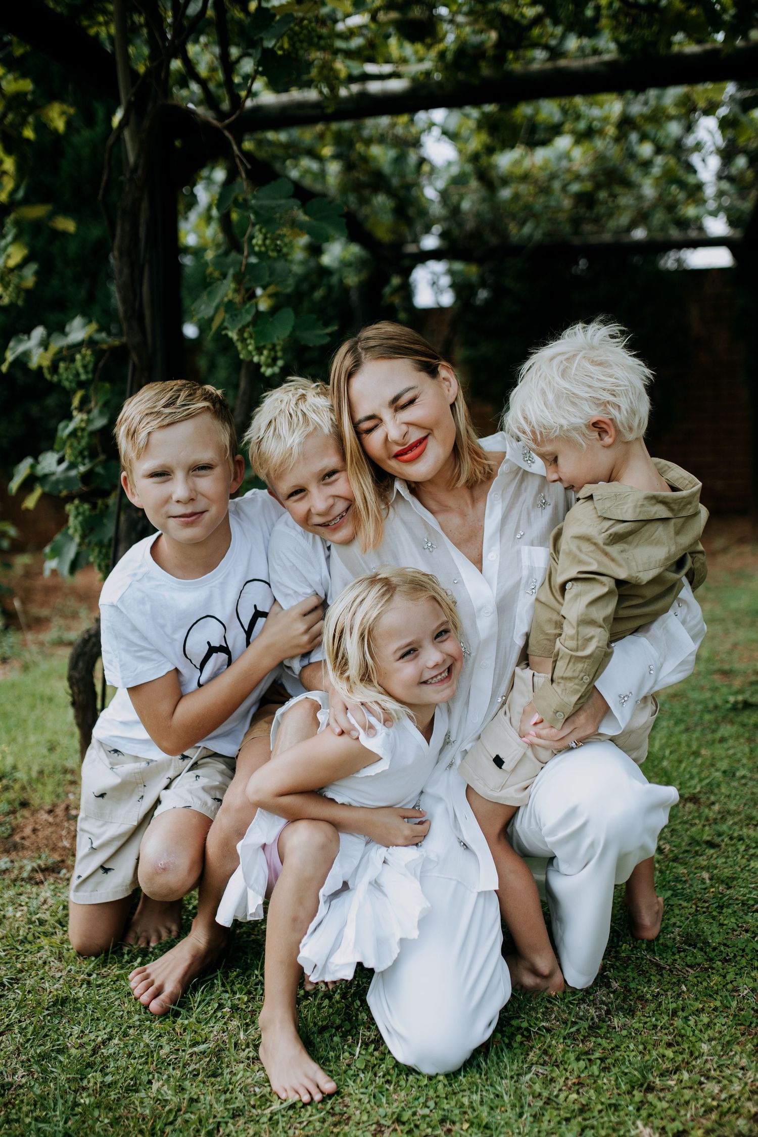 A joyful group lying on grass wearing coordinated white outfits share candid laughter together in a natural outdoor setting.