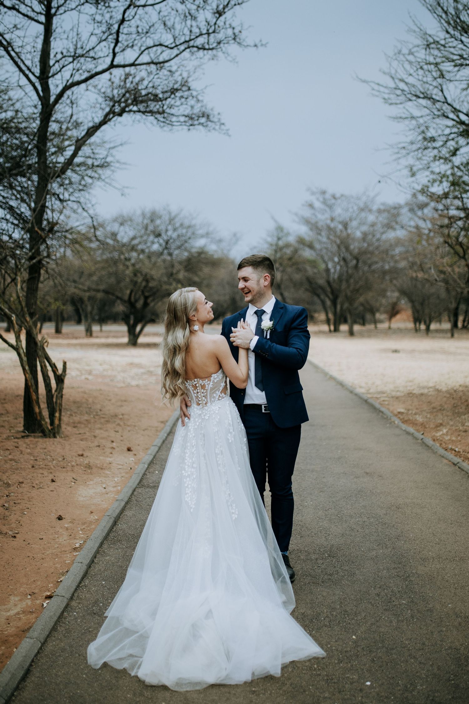 A romantic moment captured on a tree-lined path as a couple in wedding attire dance together on their special day.