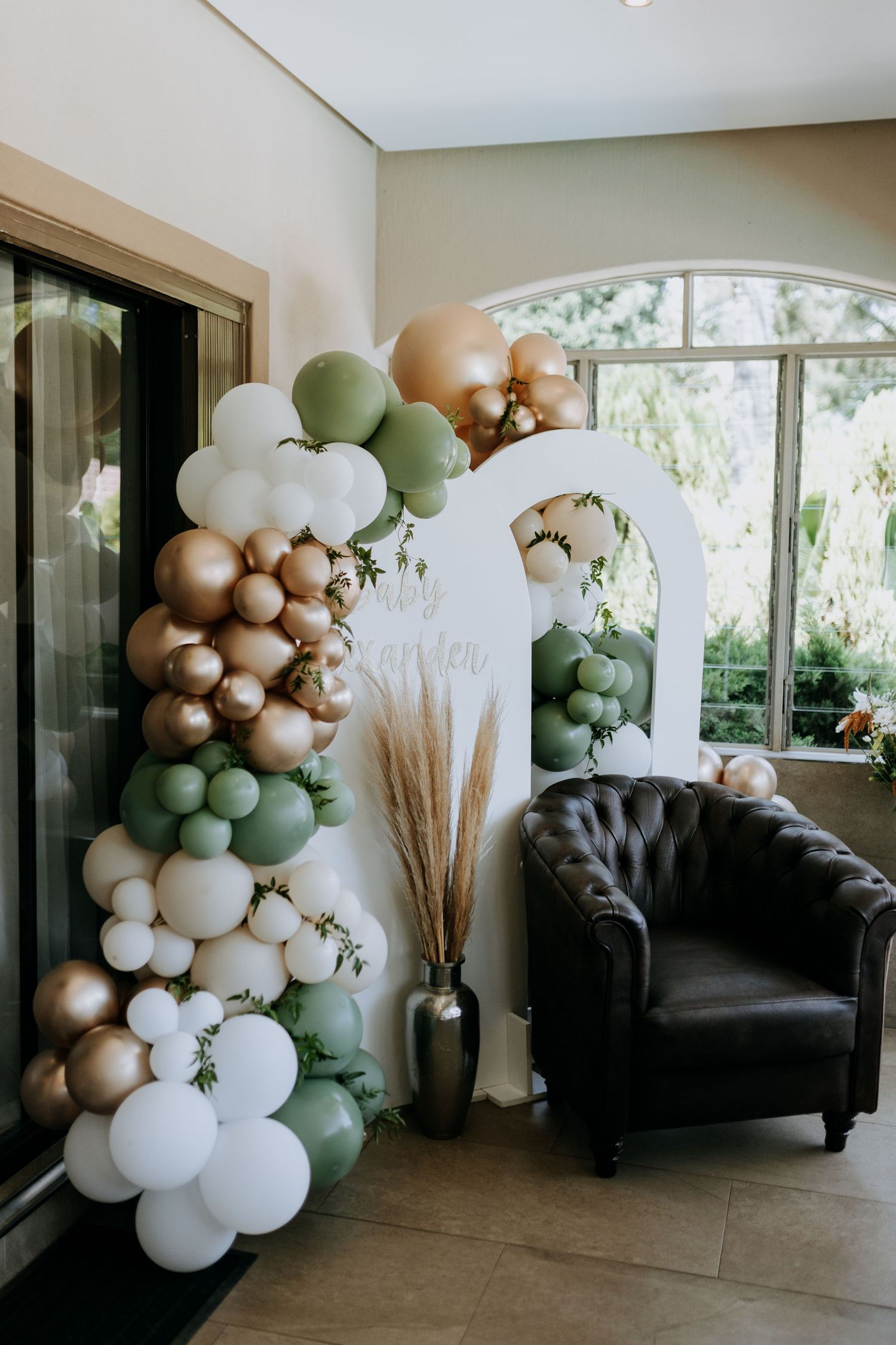 Modern event setup with an elegant balloon arch in sage green, gold and white colors with pampas grass and a black velvet chair.
