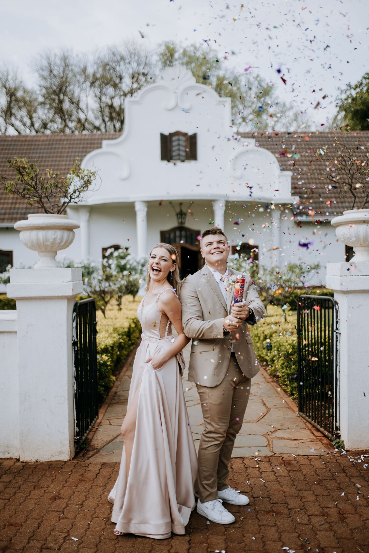 A couple celebrates their wedding day with confetti at a white colonial style entrance gate with brick pathway.
