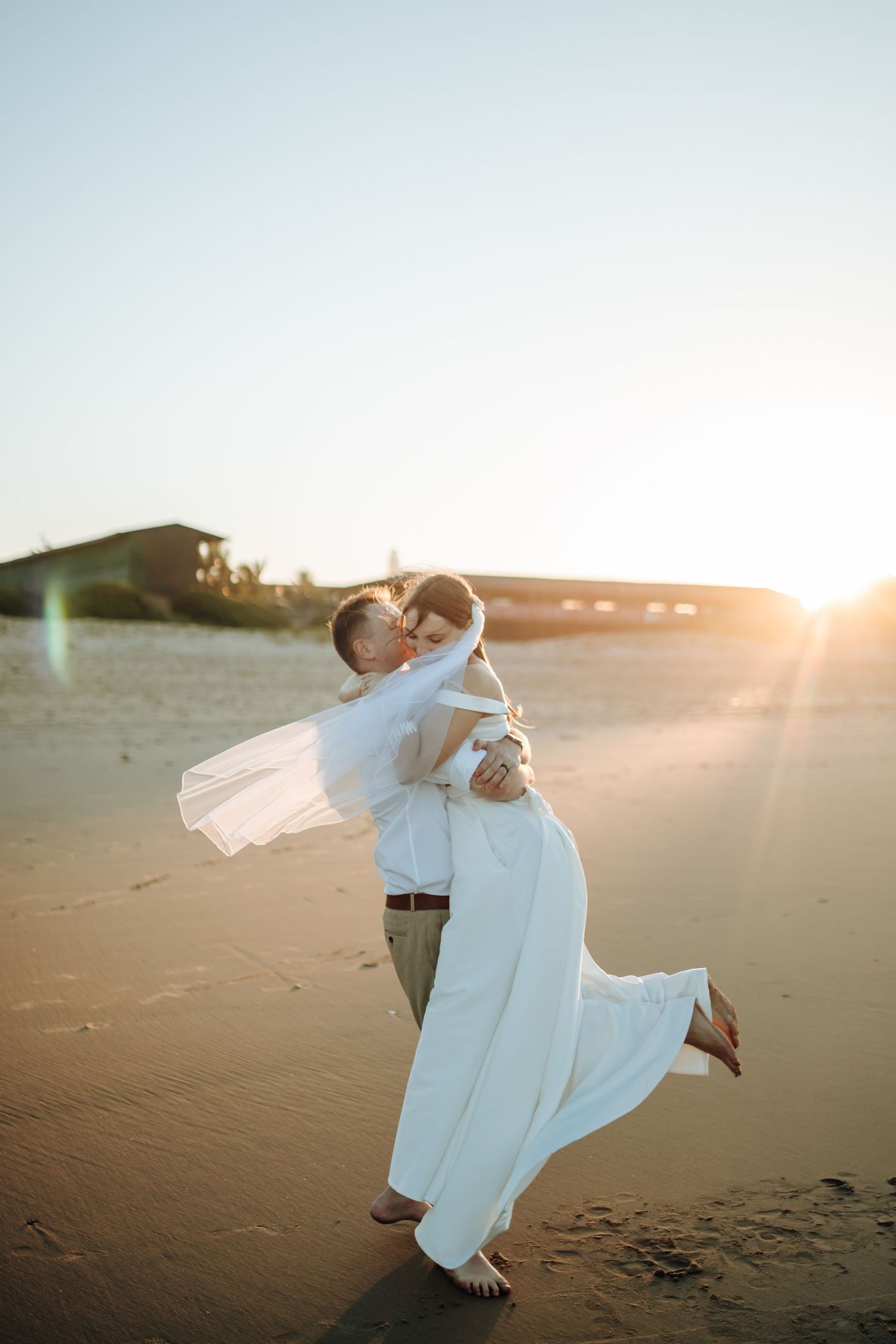 A romantic couple in white outfits dances together on a sandy beach at sunset creating dreamy silhouettes.