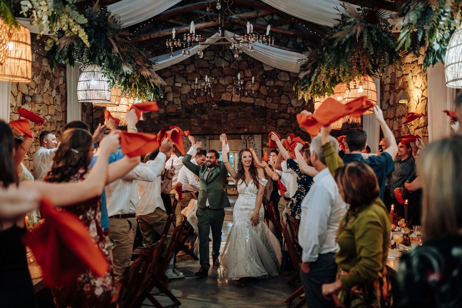 Wedding guests celebrate on a lively dance floor under large chandeliers and greenery decorations.