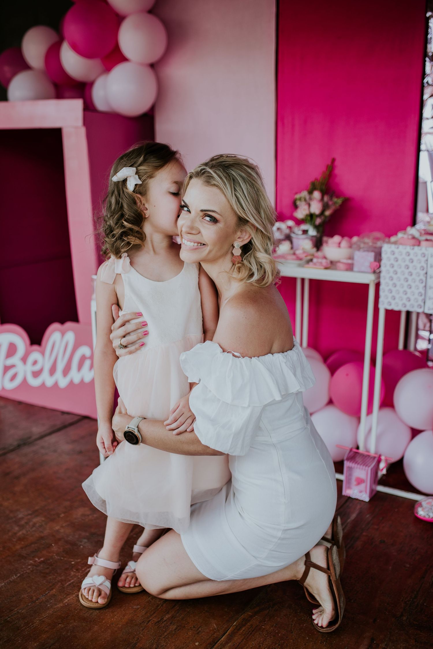 Sweet mother-daughter moment with matching white dresses and pink balloon backdrop at birthday party celebration.