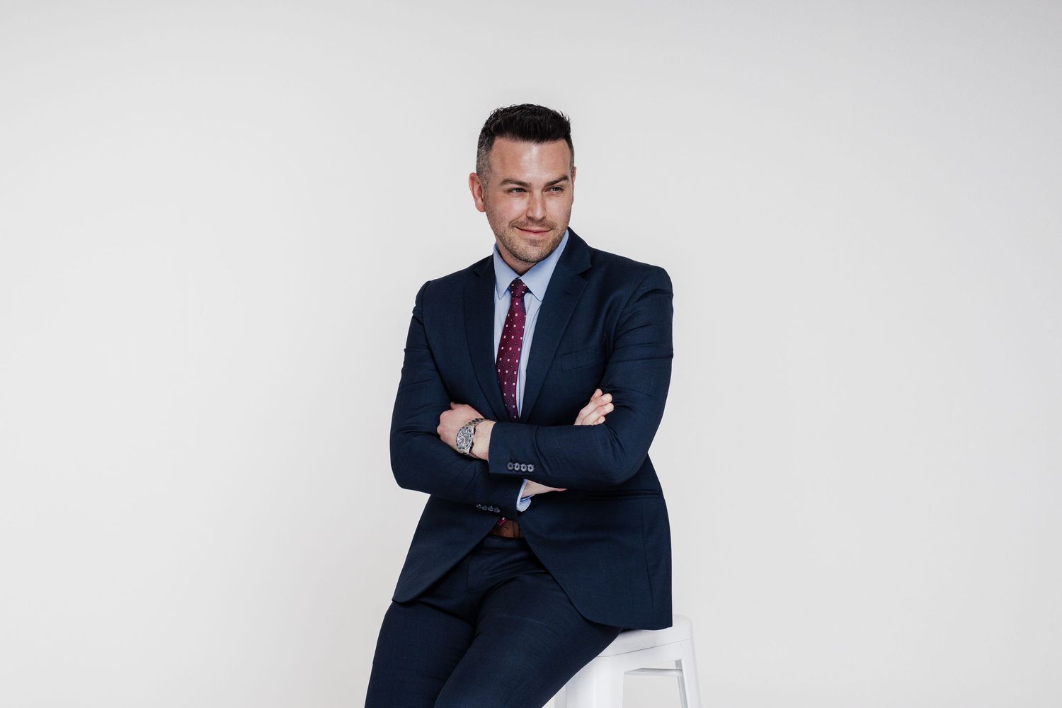 Professional wearing a navy suit and red tie sits casually on white furniture against light background.