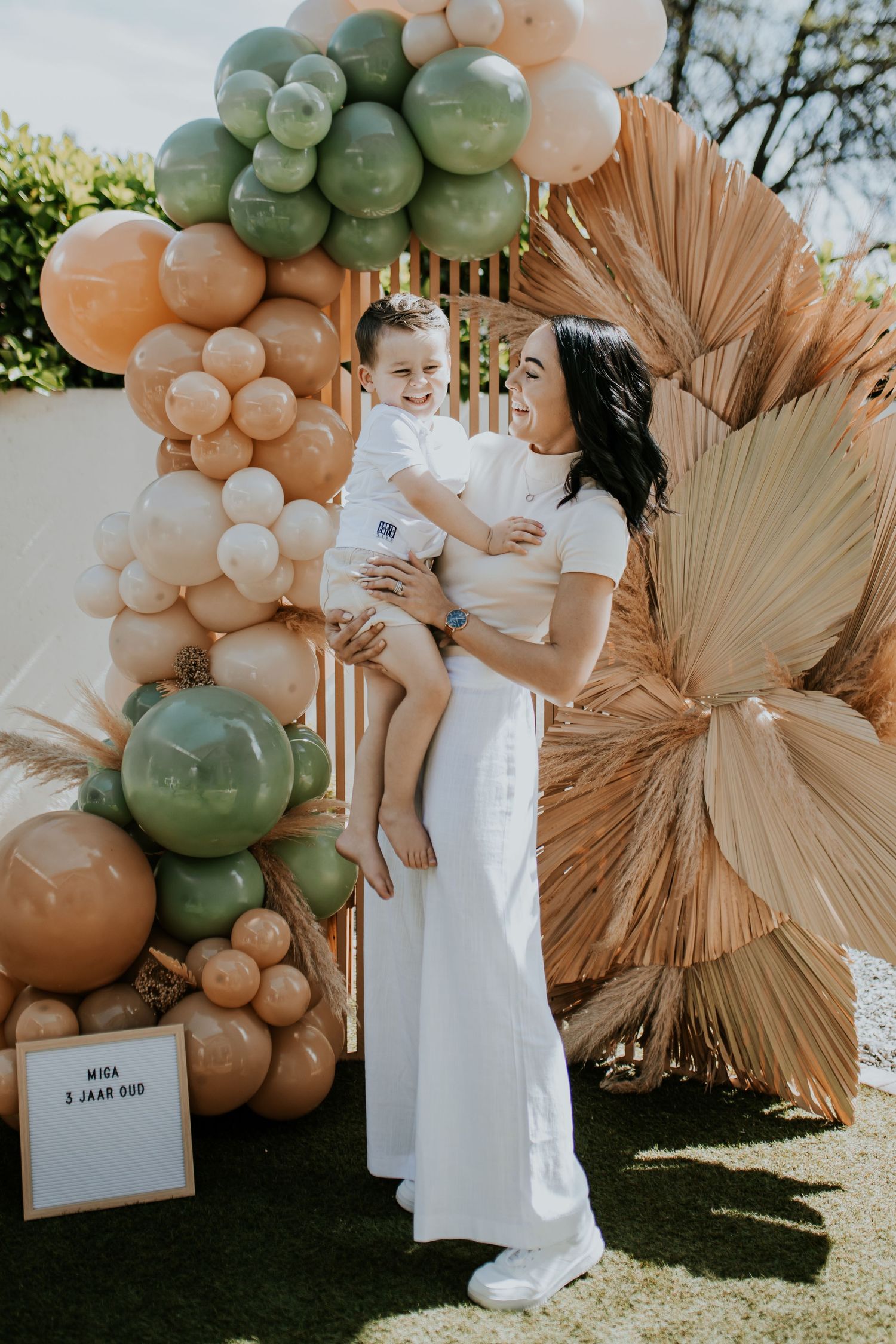 A white balloon arch installation with sage green and peach colors against dried palm fans creates a boho event backdrop.