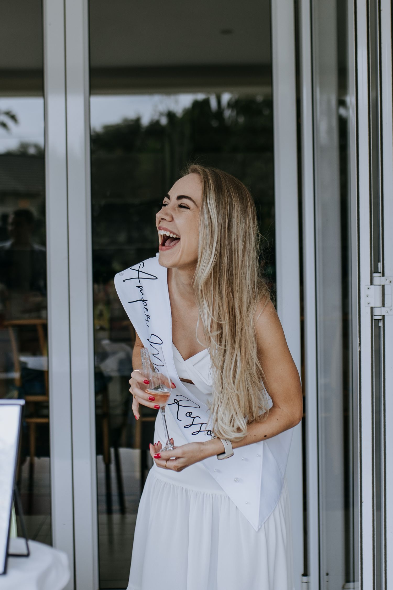 Woman in white dress laughing joyfully while holding drink cup against modern glass wall backdrop.