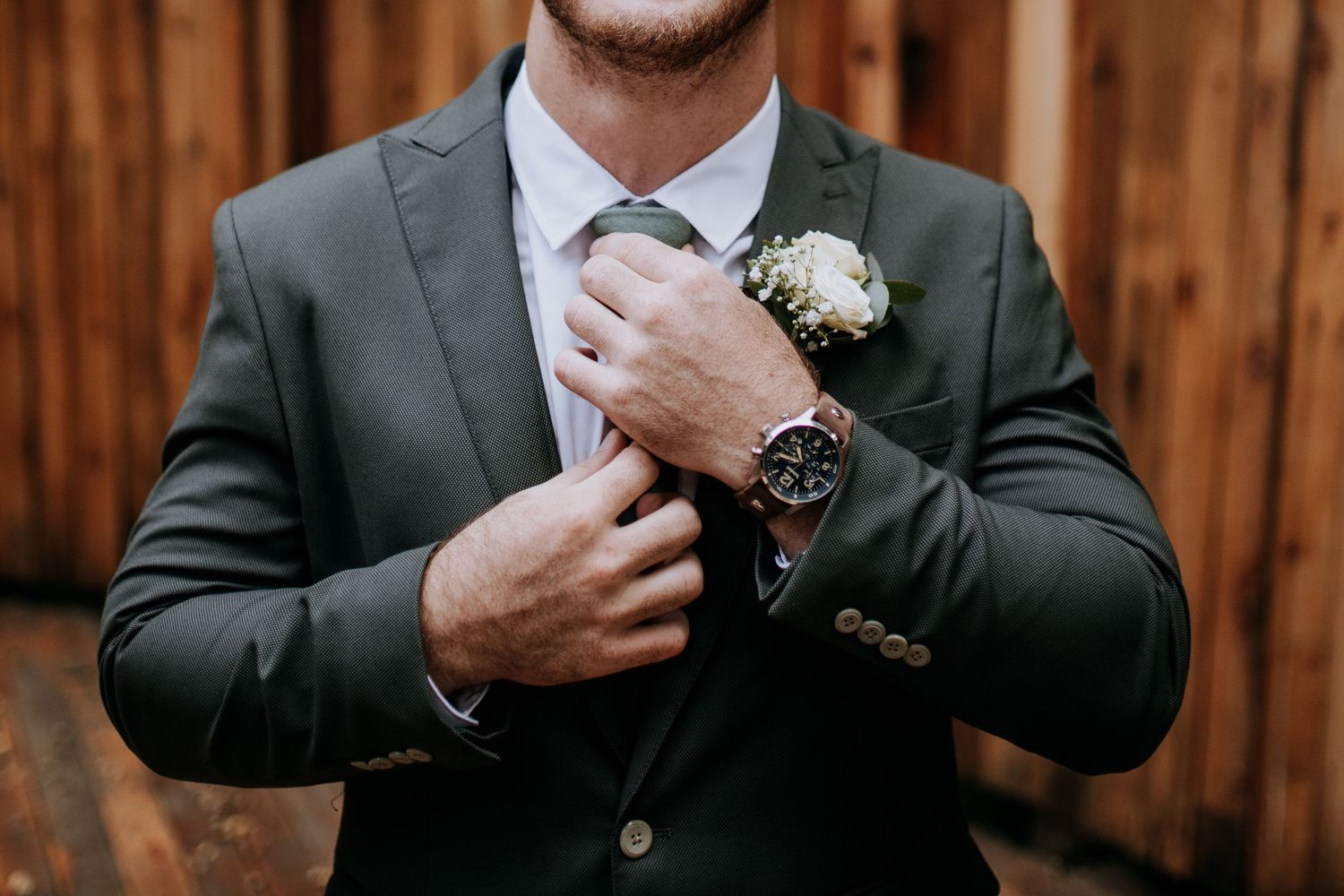 Wedding party members in dark suits make adjustments to their ties while standing against a wooden fence background.