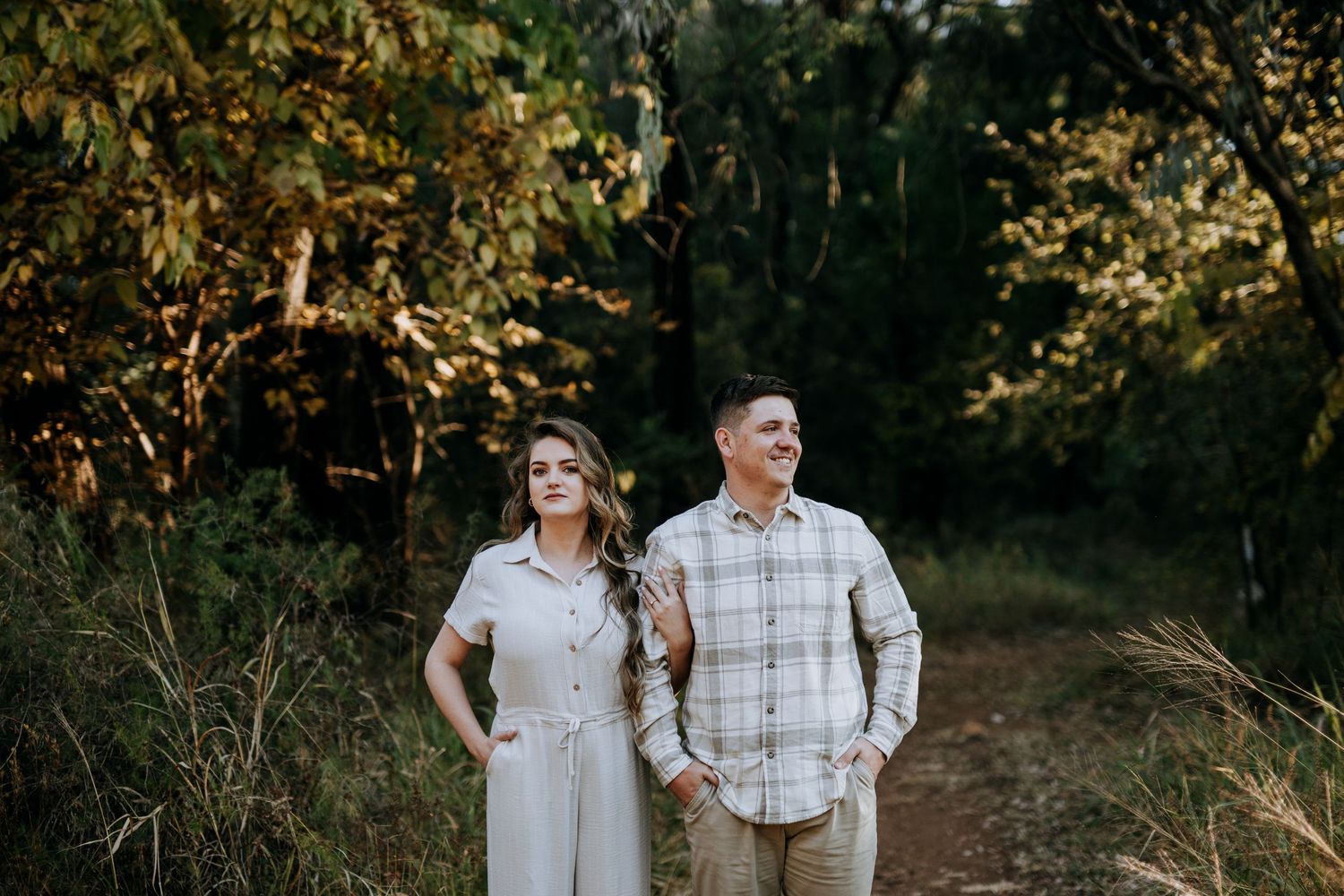 Romantic couple in light colored clothing holds hands while walking through a sunlit forest path at golden hour.