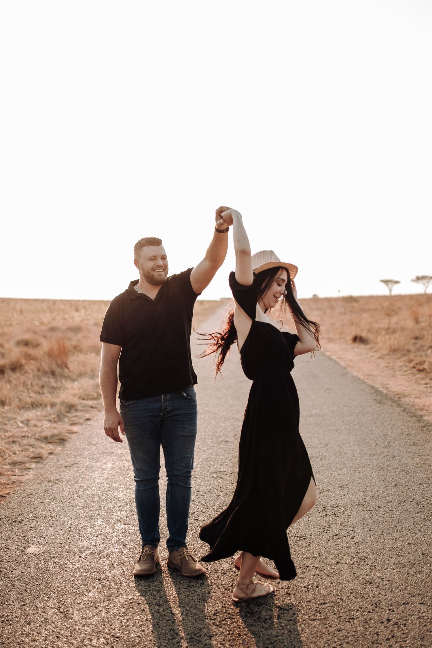 A couple dances together on a rural road during golden hour with one partner twirling in a flowing black dress.