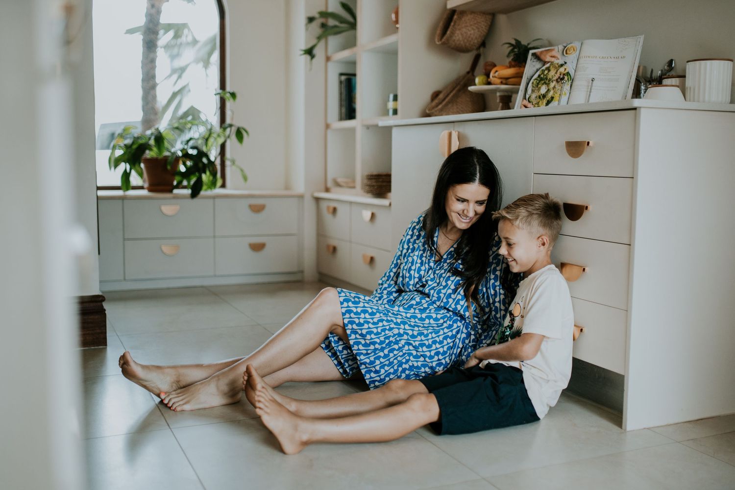 Parent and child sit together on kitchen floor reading a book near white cabinetry and potted plant by window.