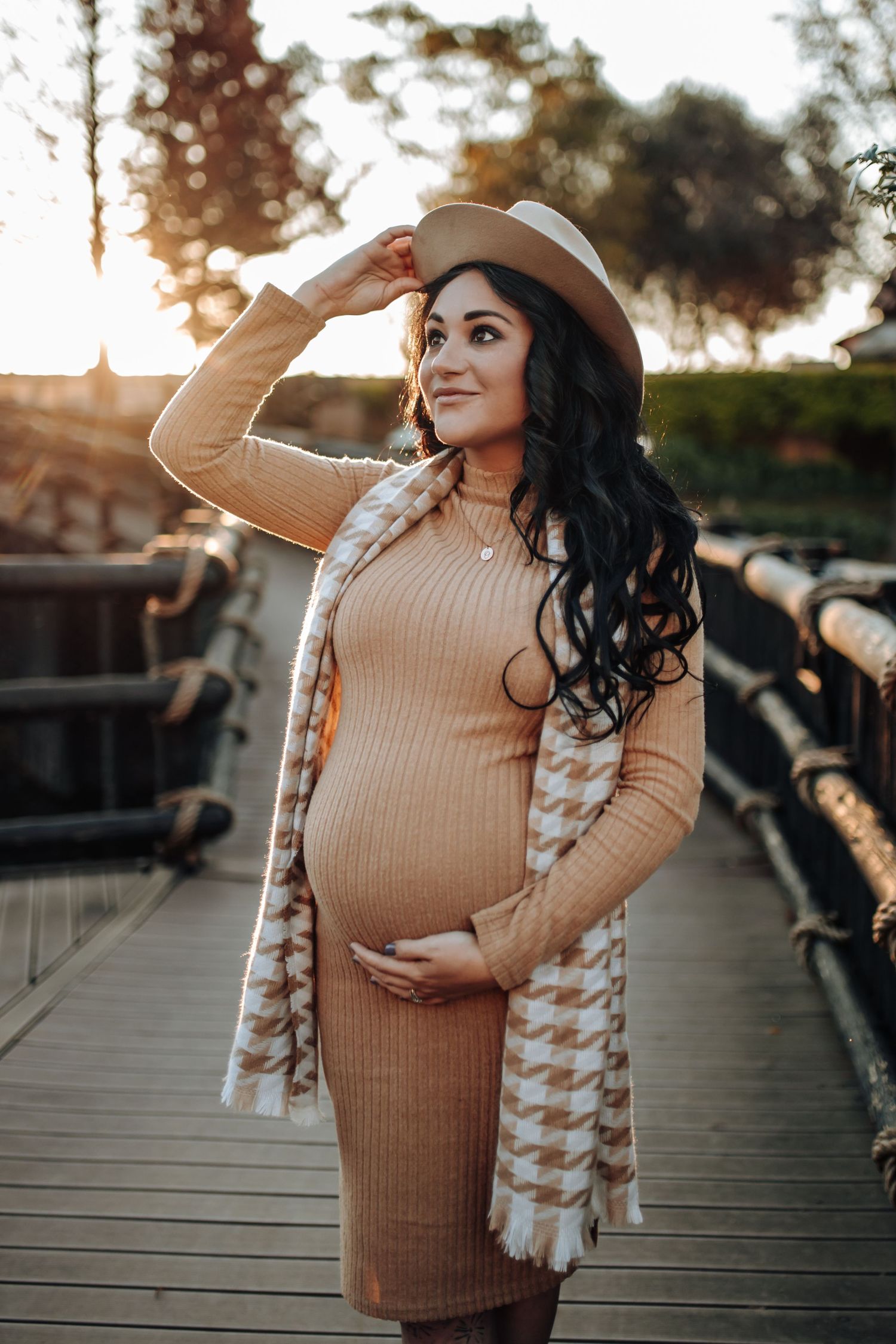 Expectant person in beige knit dress and houndstooth cardigan poses on wooden bridge at sunset while wearing wide brim hat.