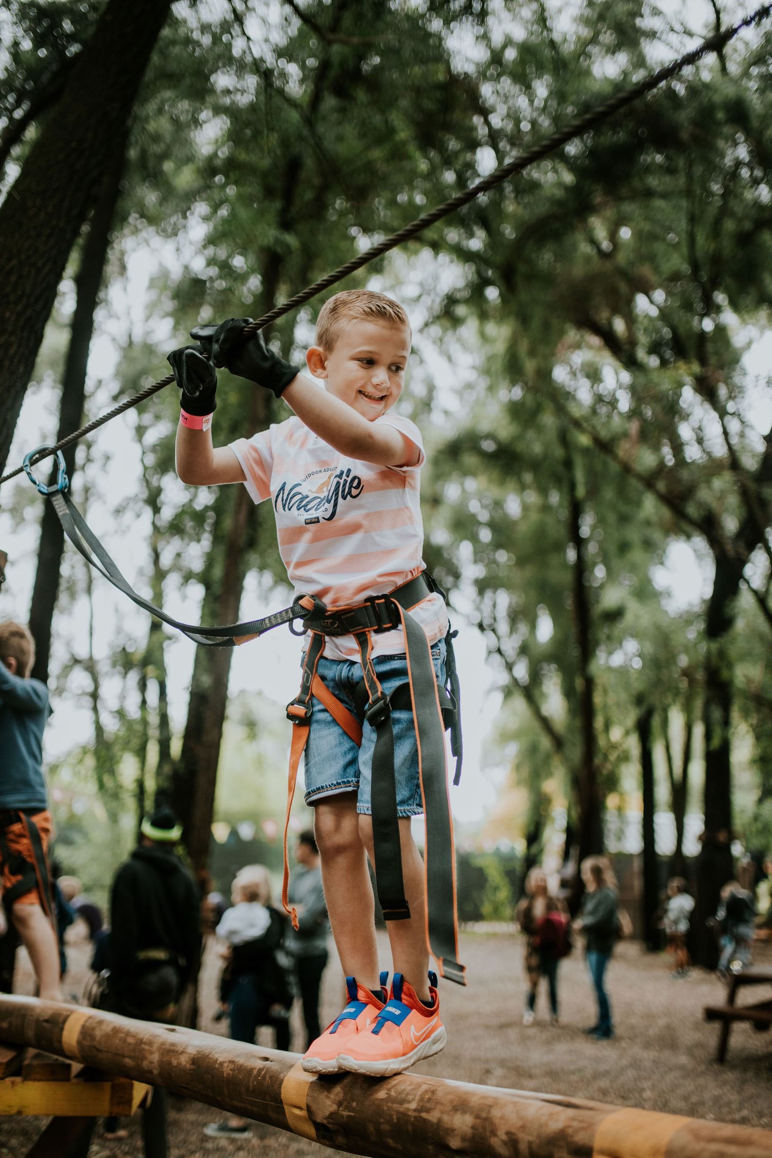 Child wearing safety harness enjoys climbing obstacle course on wooden logs in park setting.