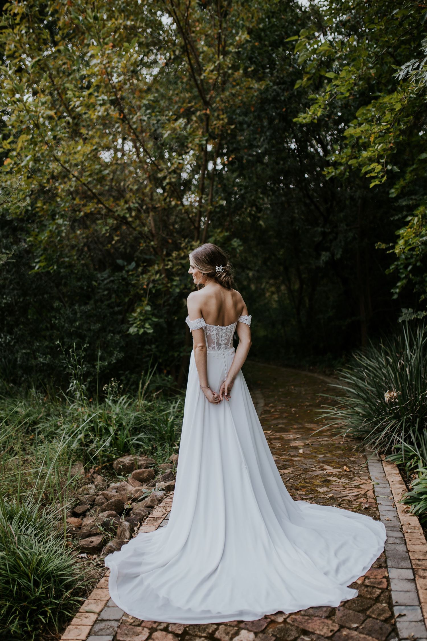 A bride in an elegant white off-shoulder wedding gown with long train stands on a stone garden path surrounded by greenery.