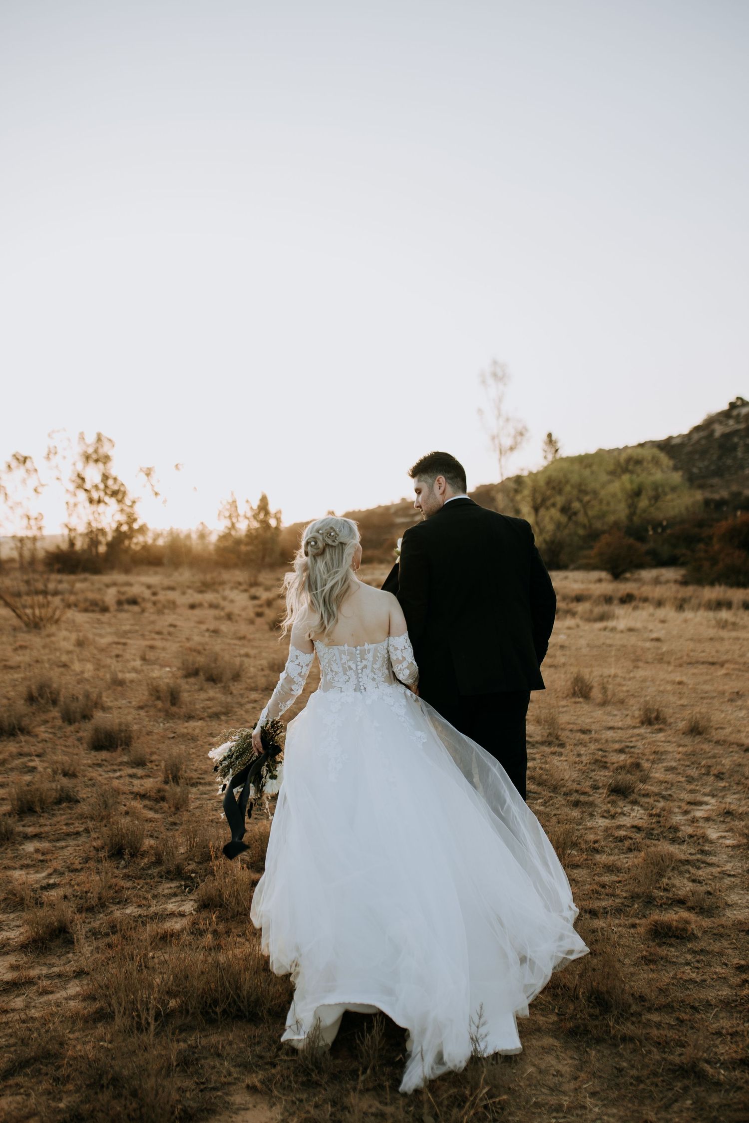 Bride and groom walk hand in hand across a desert landscape at golden hour in stylish wedding attire.