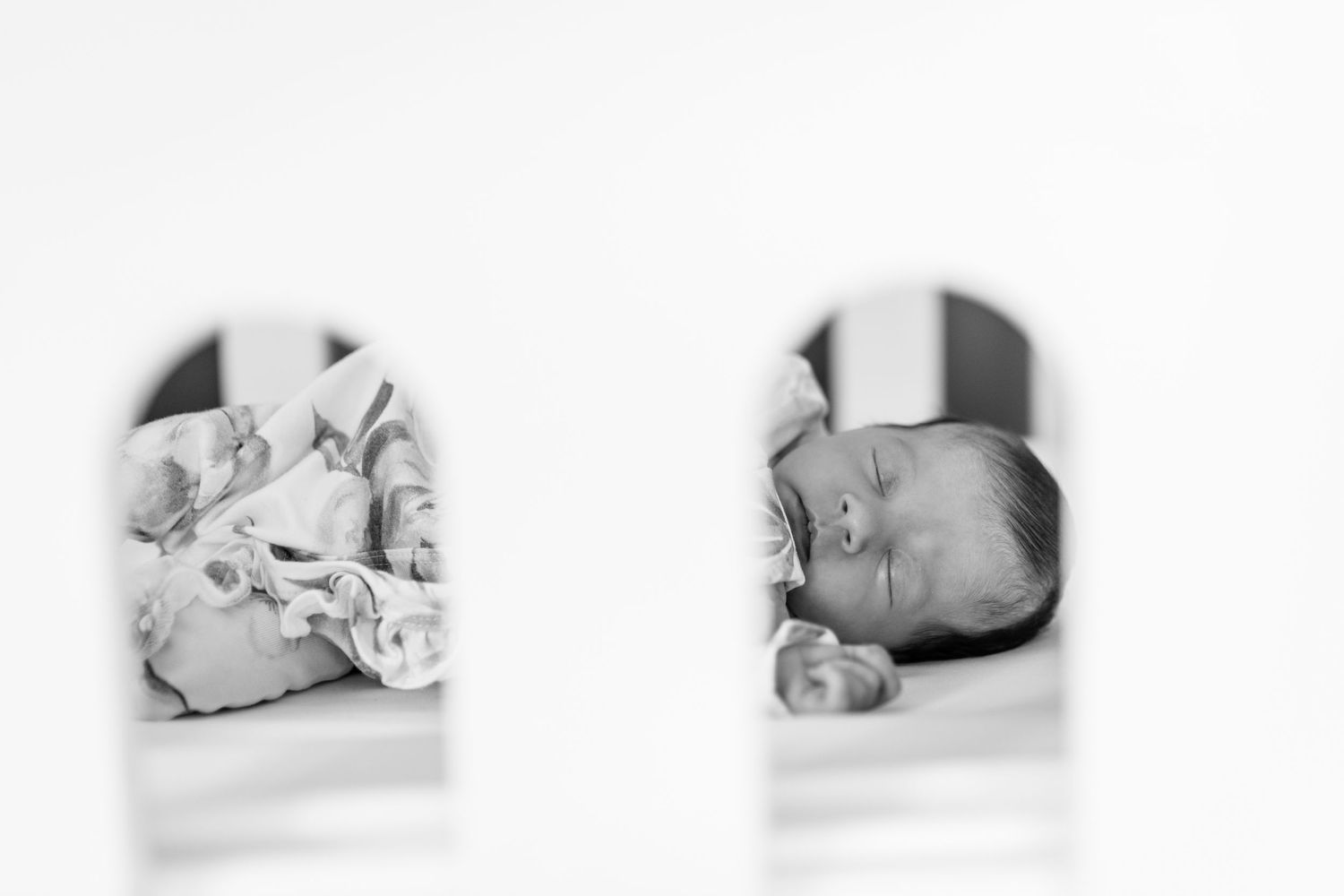 Black and white photo of a peaceful newborn baby lying in a white crib, viewed through crib slats.