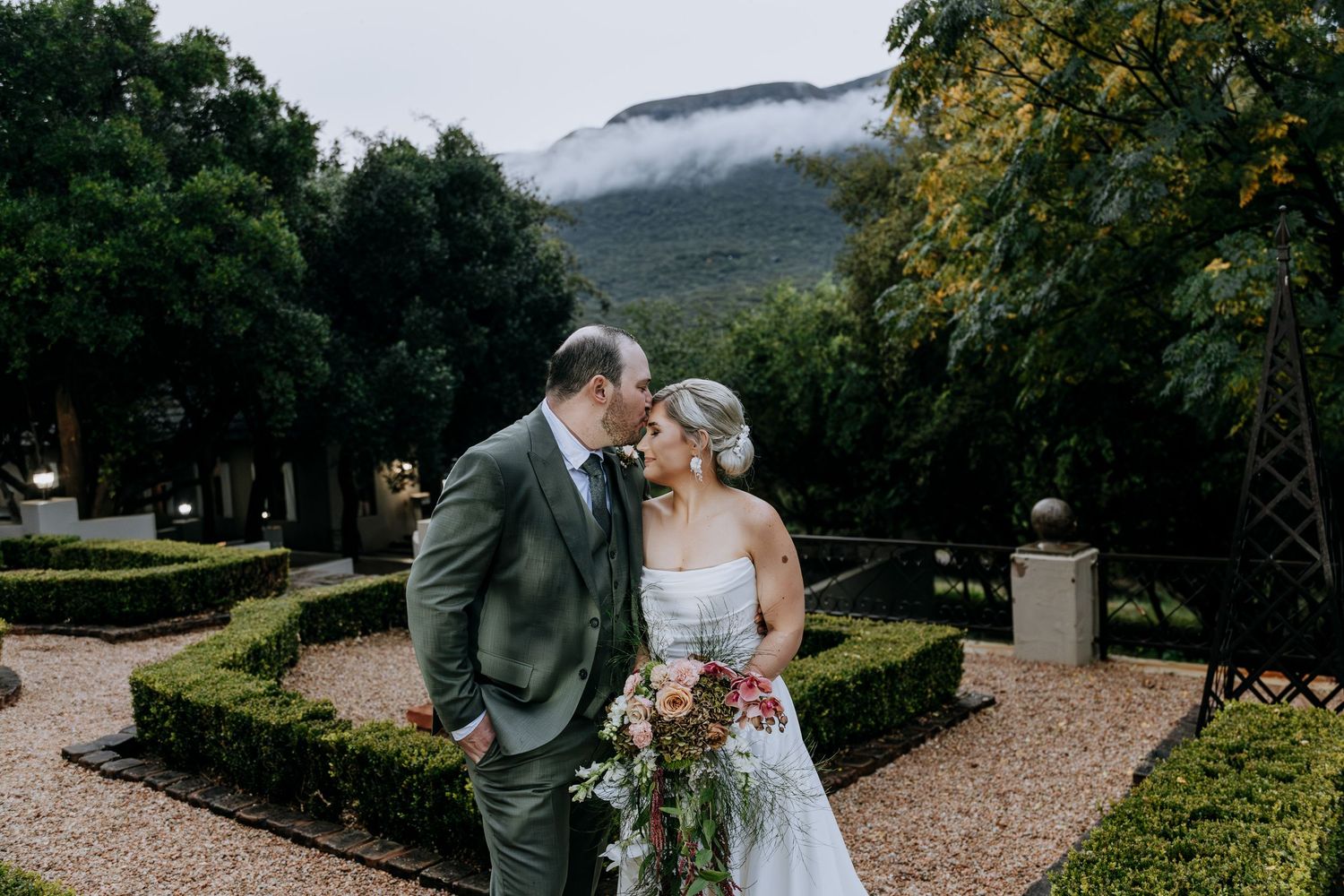 A couple shares a romantic moment together in a scenic garden with mountain views and lush greenery in the background.