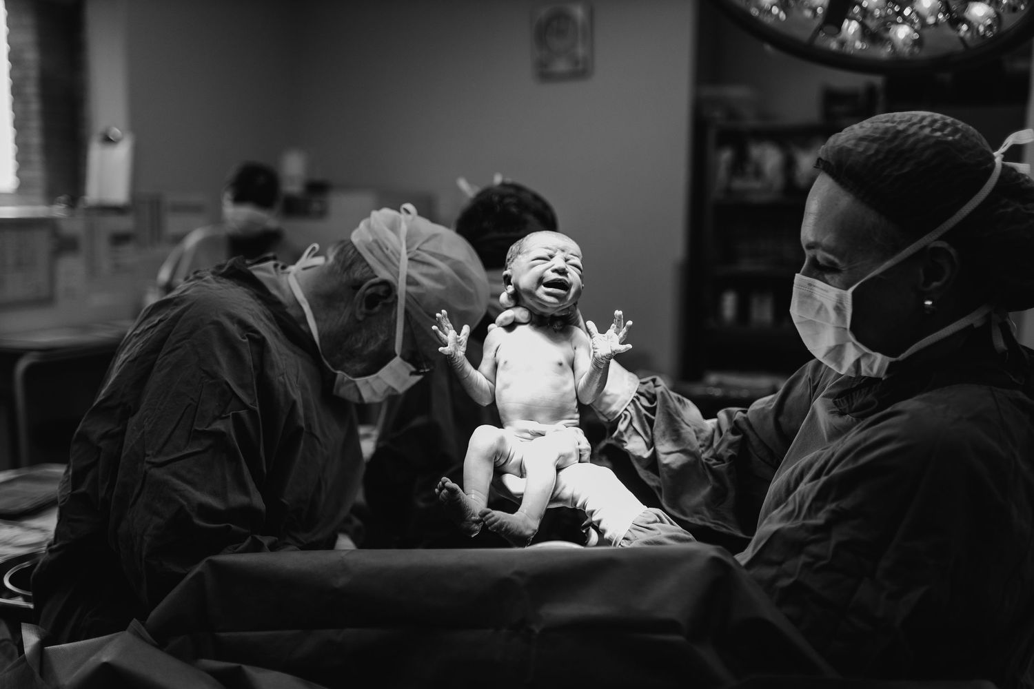 Newborn baby crying with arms raised moments after birth in operating room with medical staff in scrubs and masks.