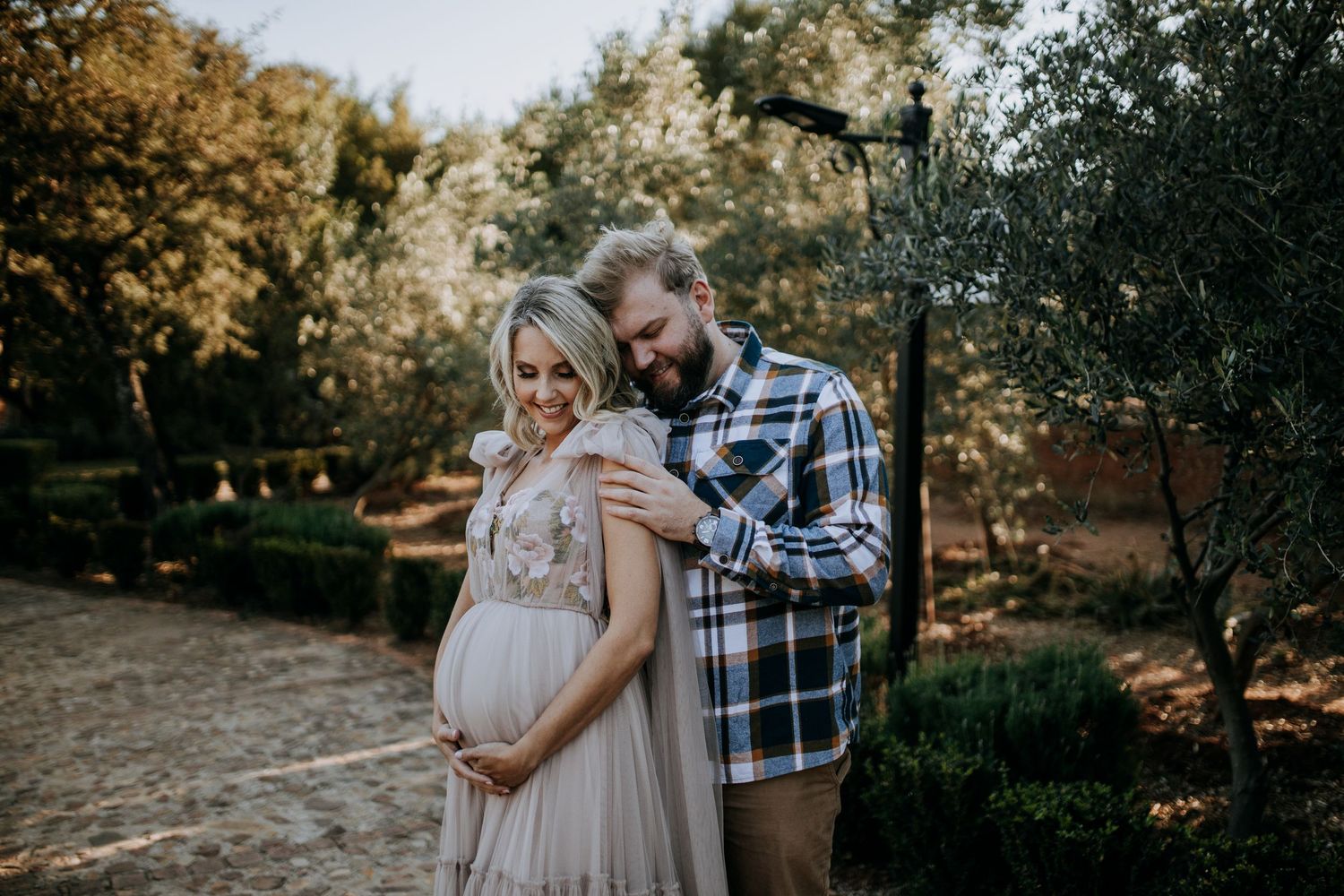 Couple sharing a tender maternity moment outdoors in a garden, woman in flowing gown cradling baby bump.