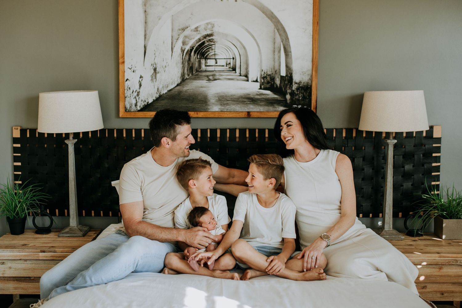 Happy family of five sitting together on a bed, smiling and bonding in a cozy bedroom with neutral decor.