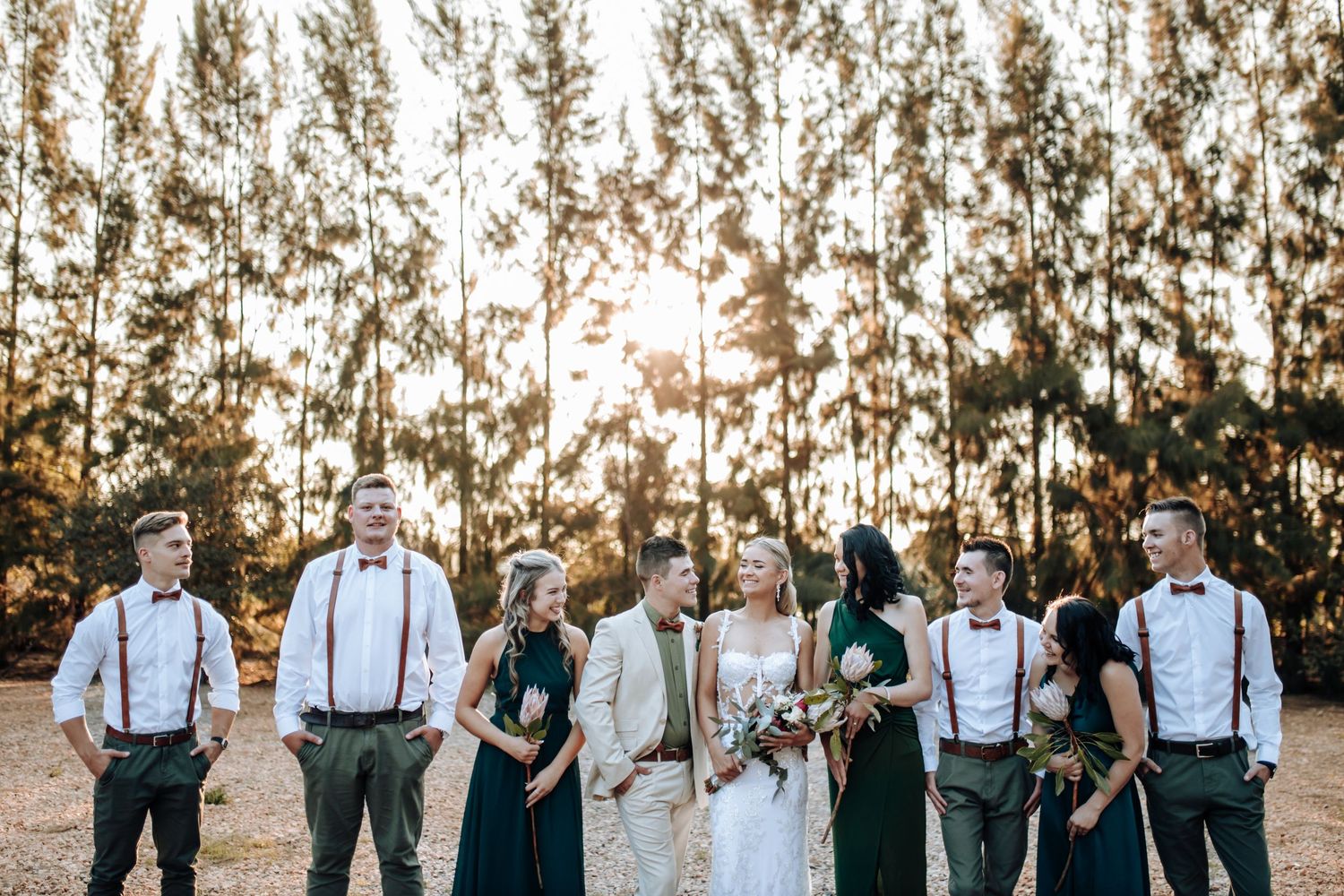 Bridal party in rustic attire with suspenders poses together at sunset for group wedding photos in countryside setting.