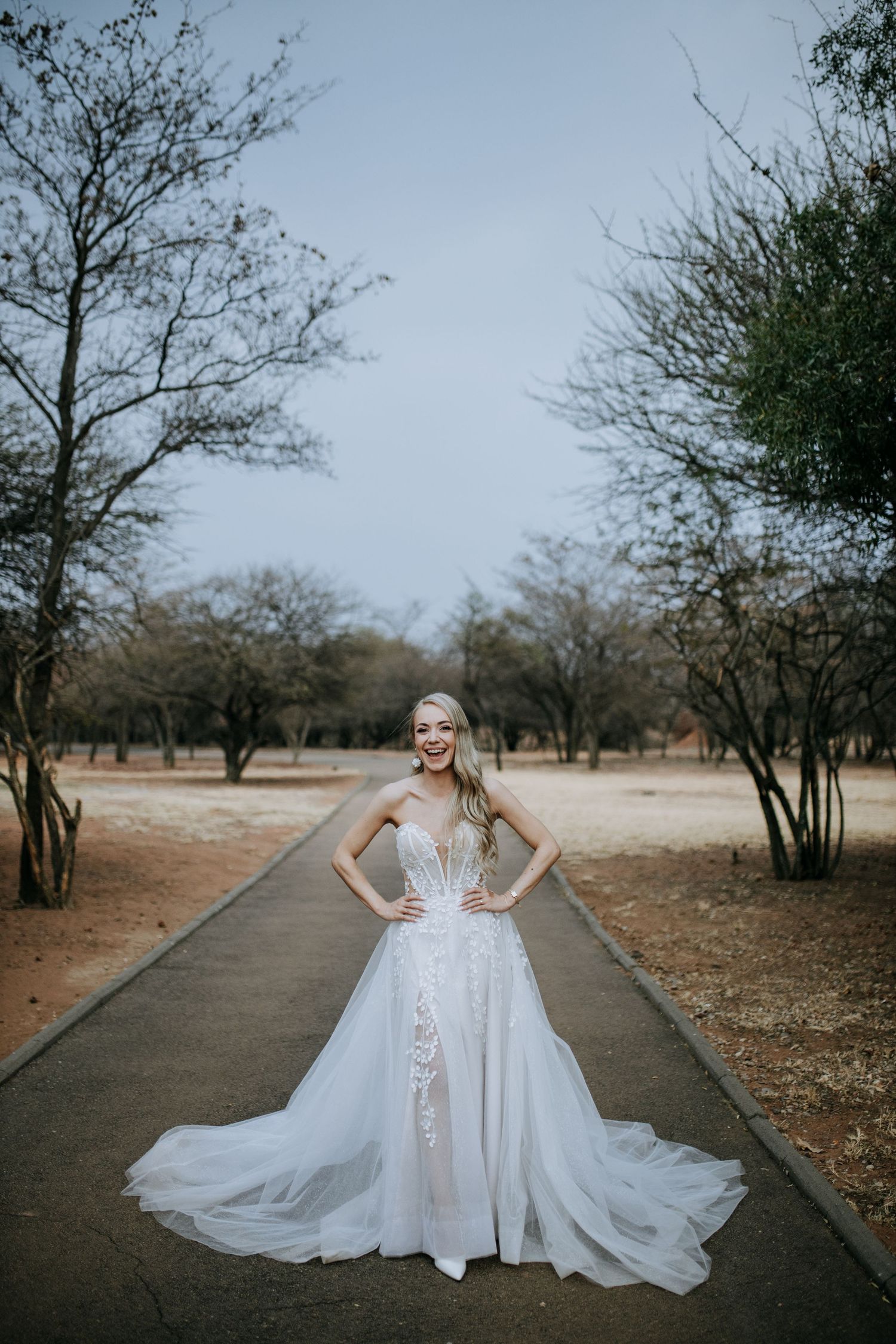 White wedding dress with flowing train and lace details photographed in moody outdoor setting with bare trees.