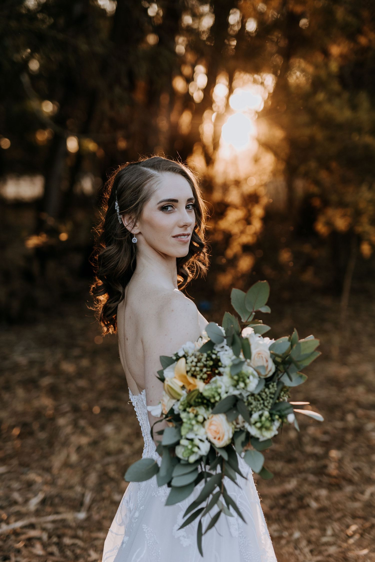 A bride in a white strapless wedding gown holds a peach and white floral bouquet at sunset in a wooded setting.