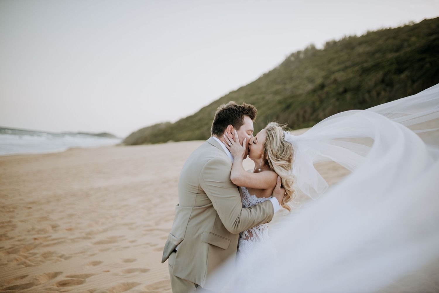 A couple embraces on a sandy beach at sunset with hills in the background.